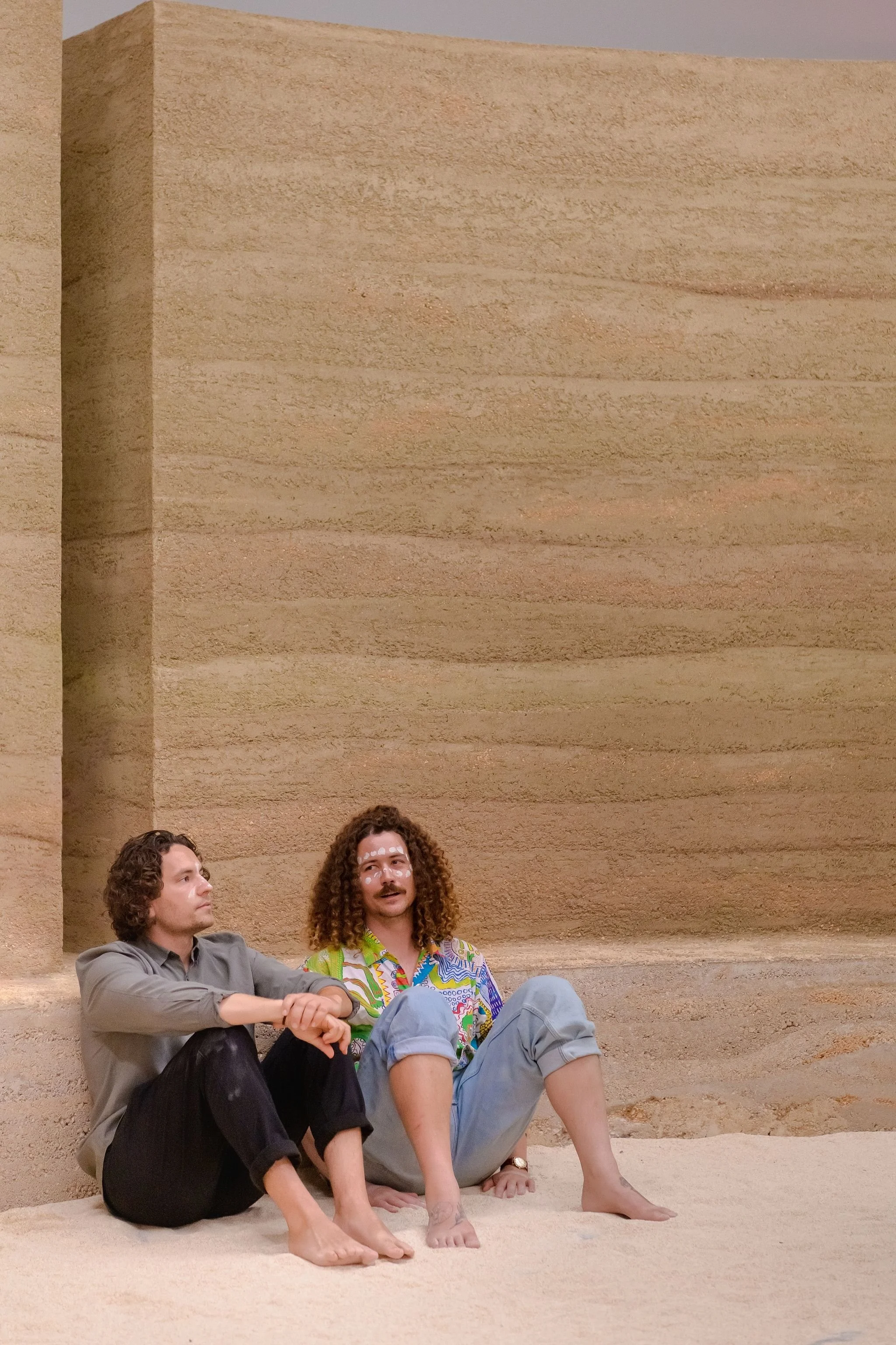 Two men sitting barefoot on the sand against the rammed earth wall, one wearing white ochre markings