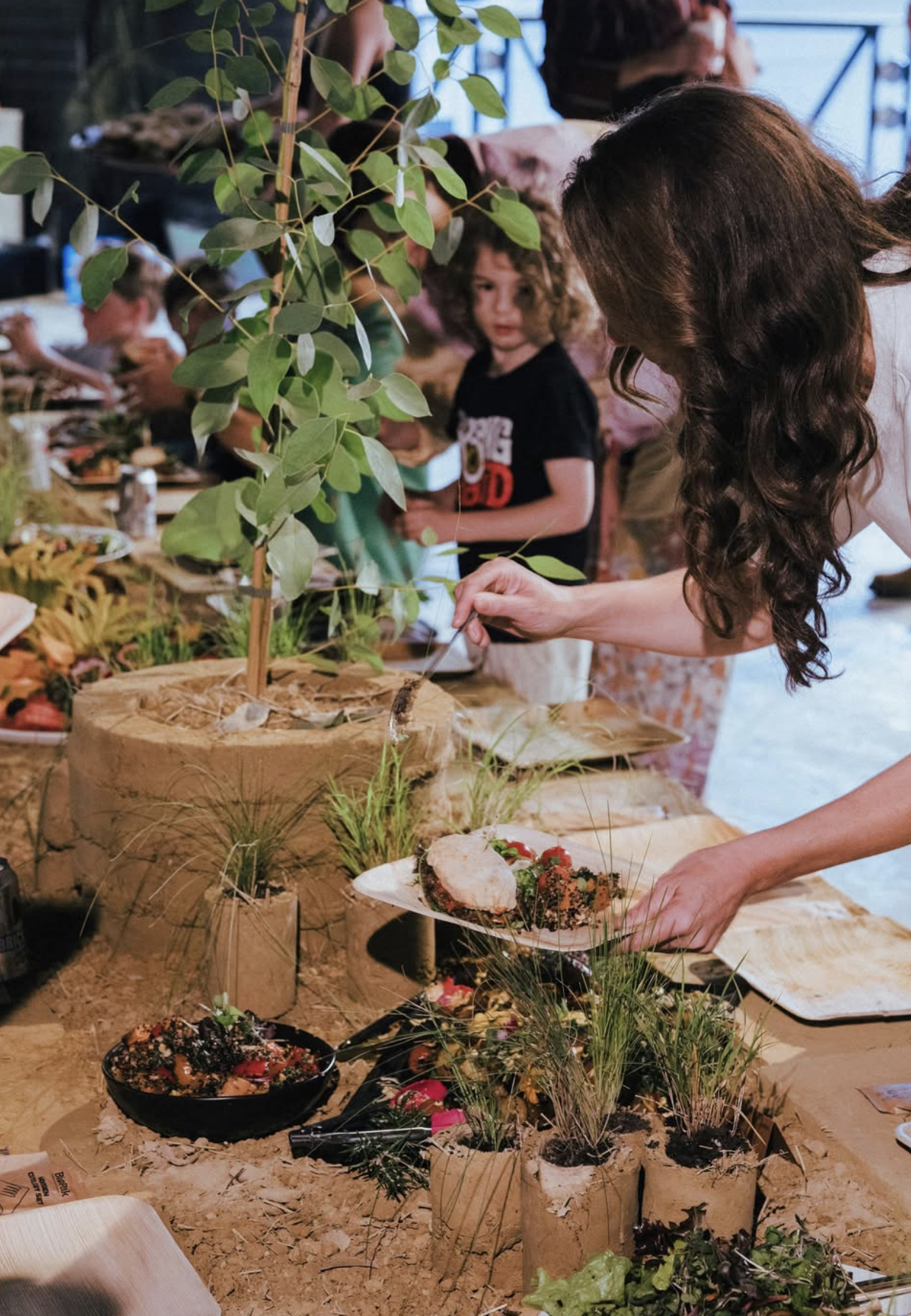 Guests serving themselves from a table of food arranged among potted grasses and a young gum tree