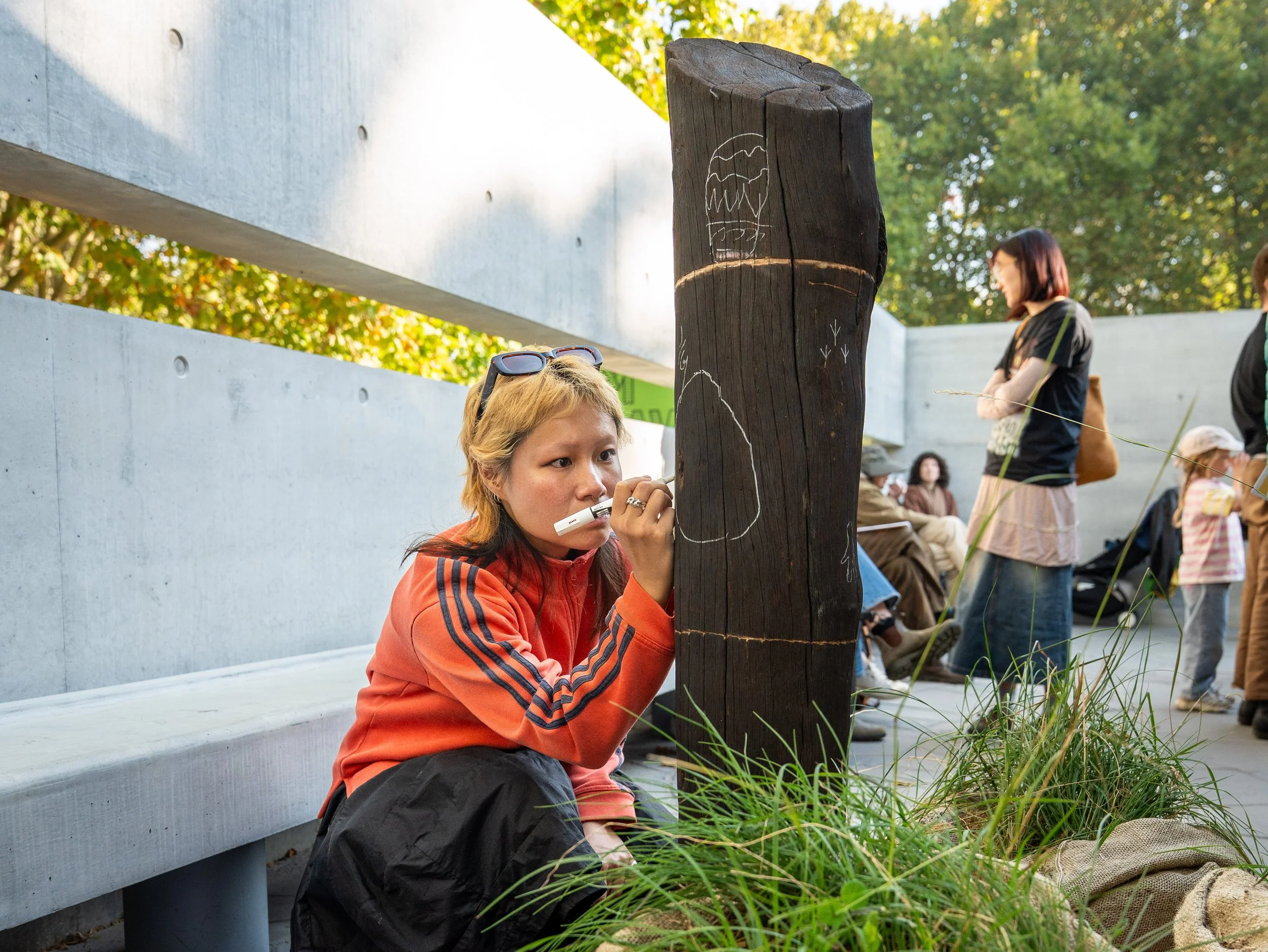 A visitor in an orange jacket drawing on a charred timber post with a white marker