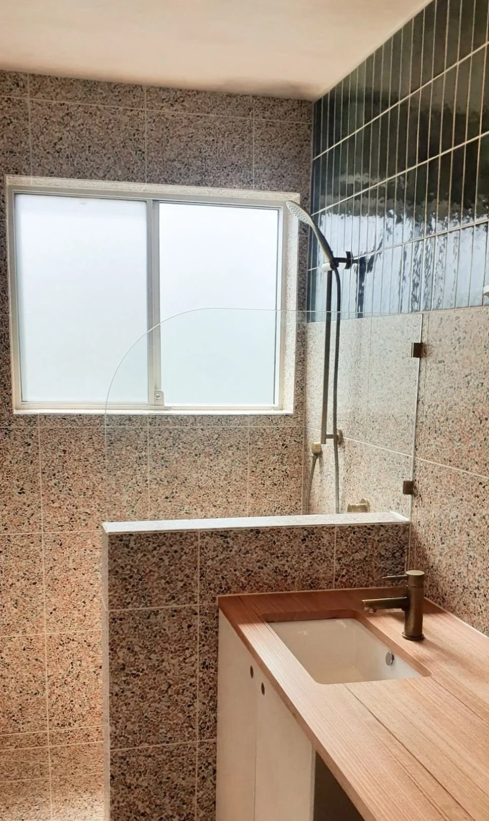 Bathroom with speckled tiles, a frameless glass shower screen and a timber vanity beneath the window