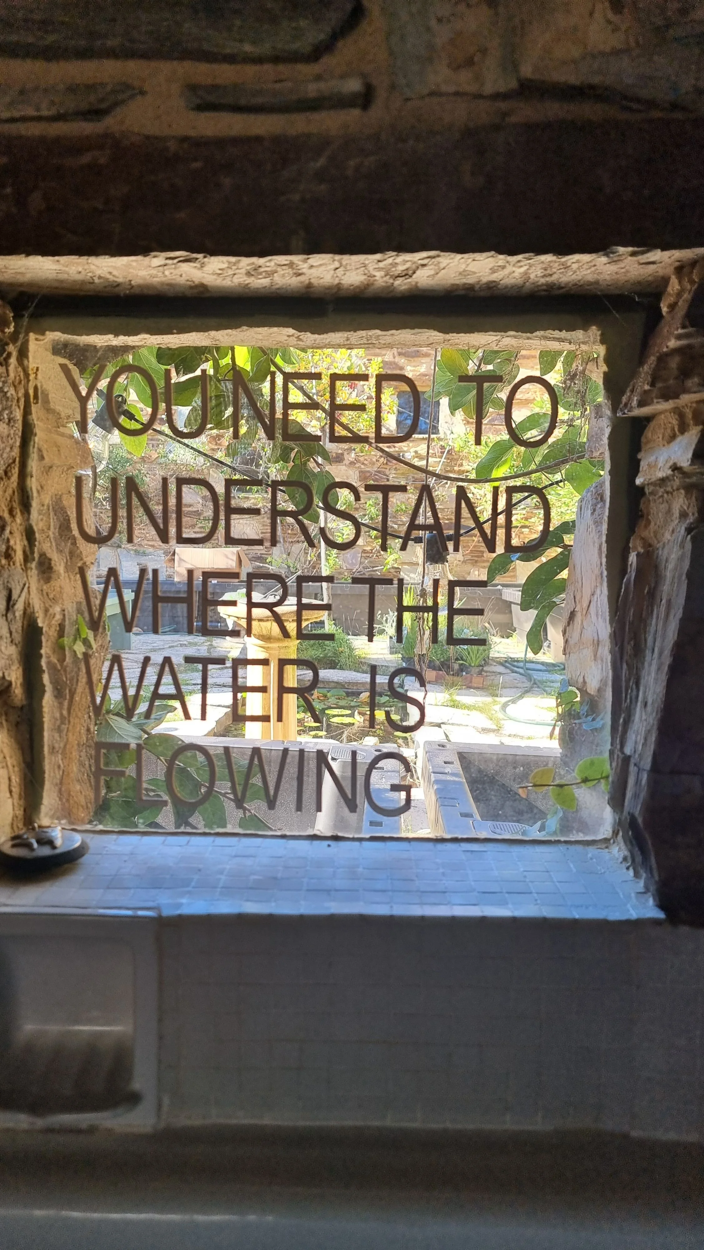 Window lettering reading "You need to understand where the water is flowing", looking through a stone-framed window into a leafy courtyard