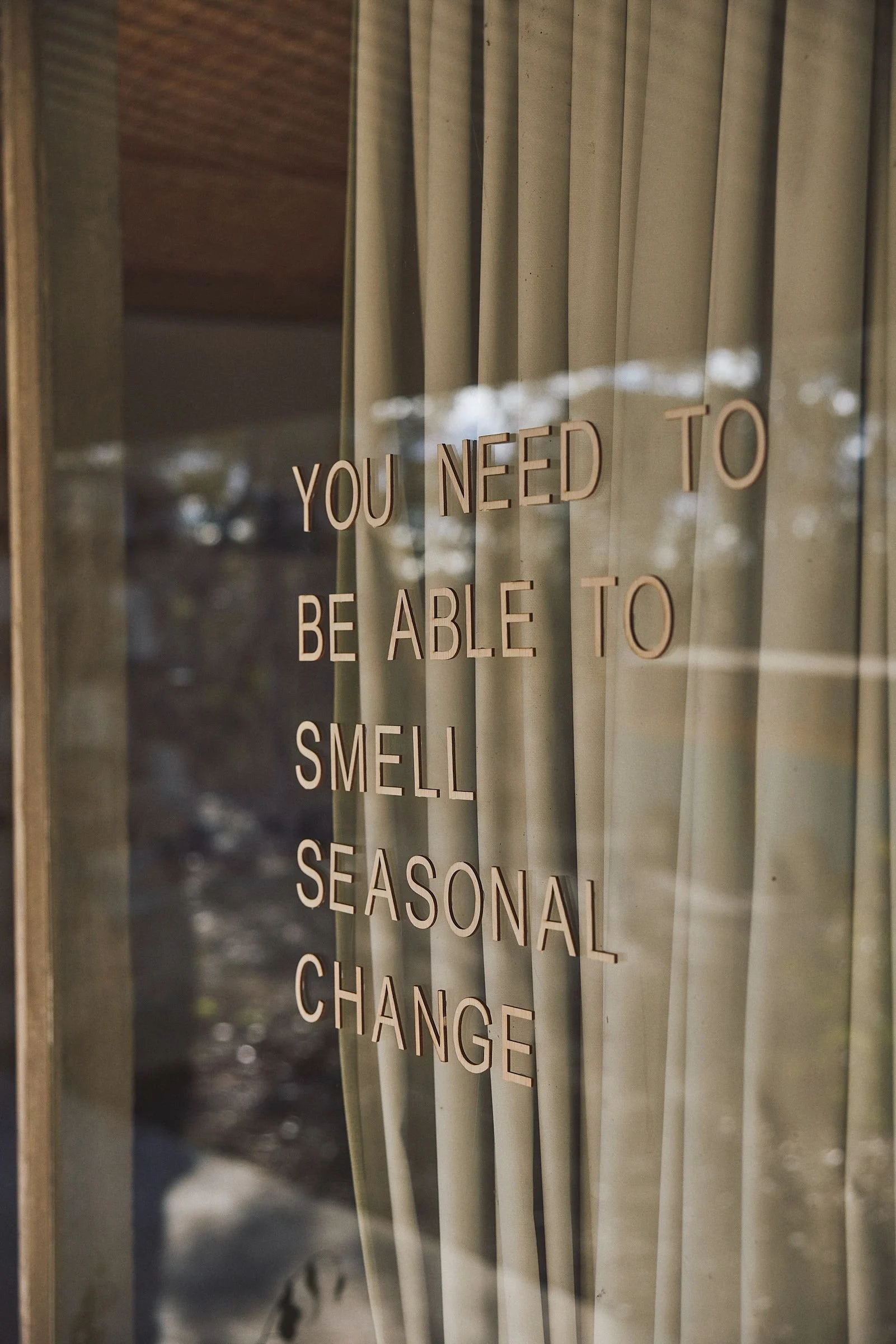 Window lettering reading "You need to be able to smell seasonal change" over linen curtains