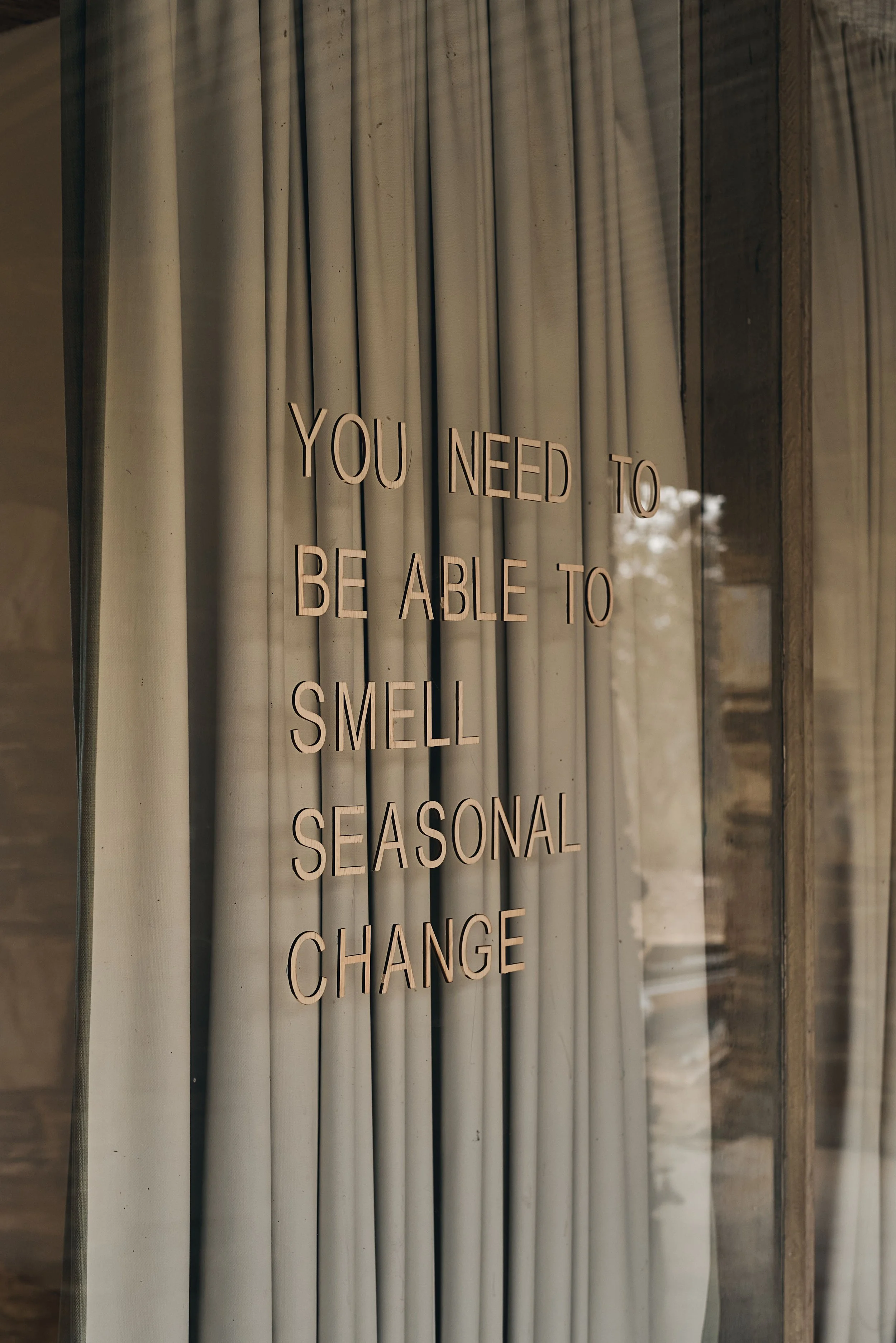 The same "You need to be able to smell seasonal change" lettering seen closer, in soft light against the curtains