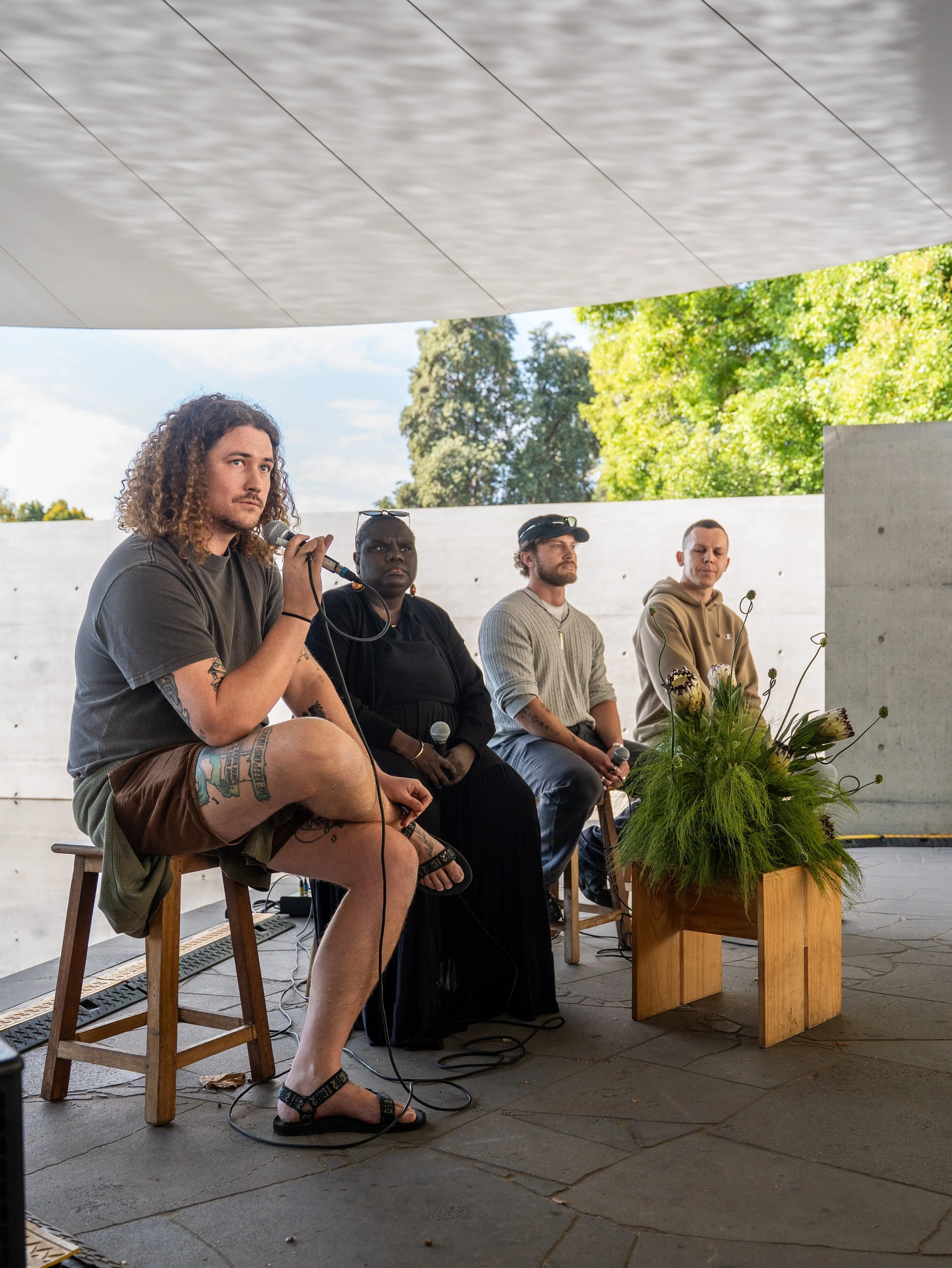 Bradley Kerr speaking into a microphone beside three panellists beneath the MPavilion canopy
