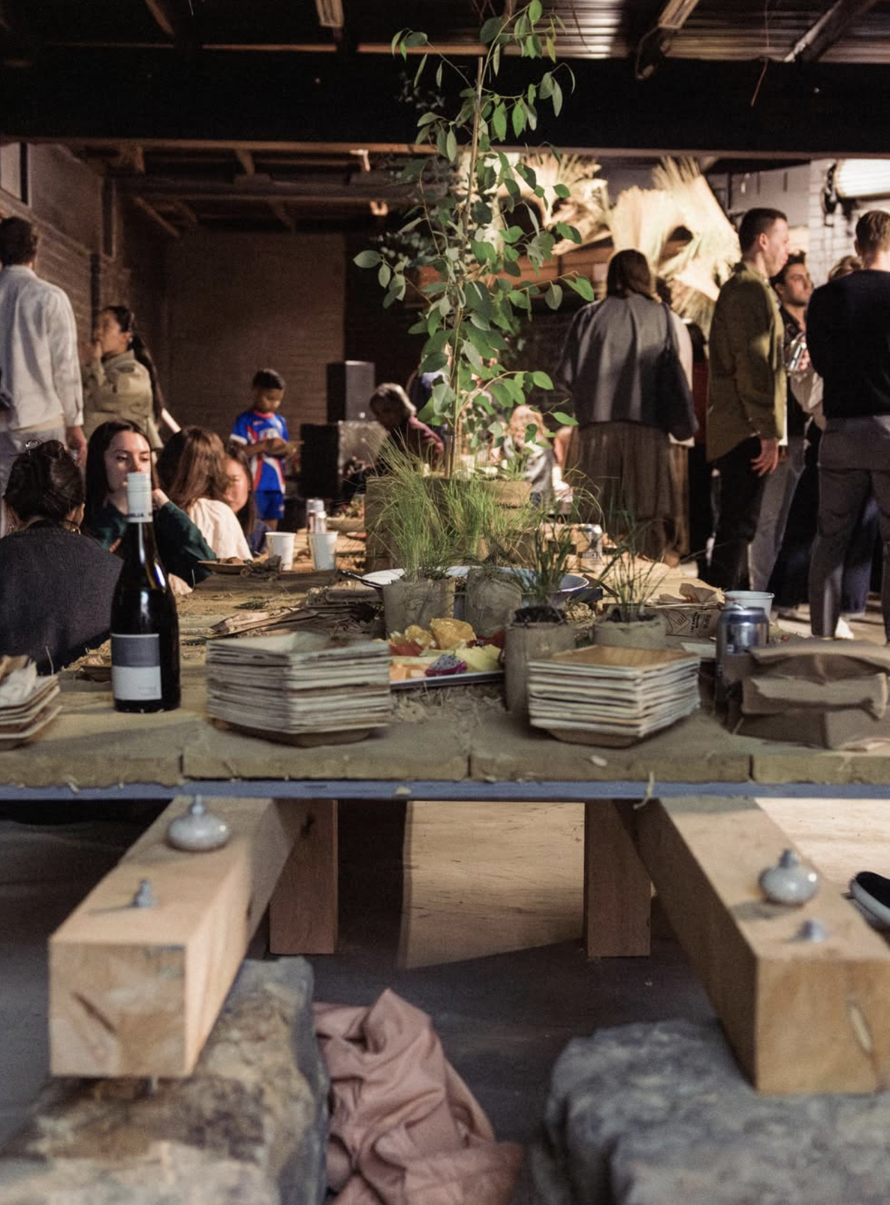 But First, Country at Melbourne Design Week 2026: a long communal table set with stacked handmade plates, food and potted native plants, as guests gather in a warehouse space