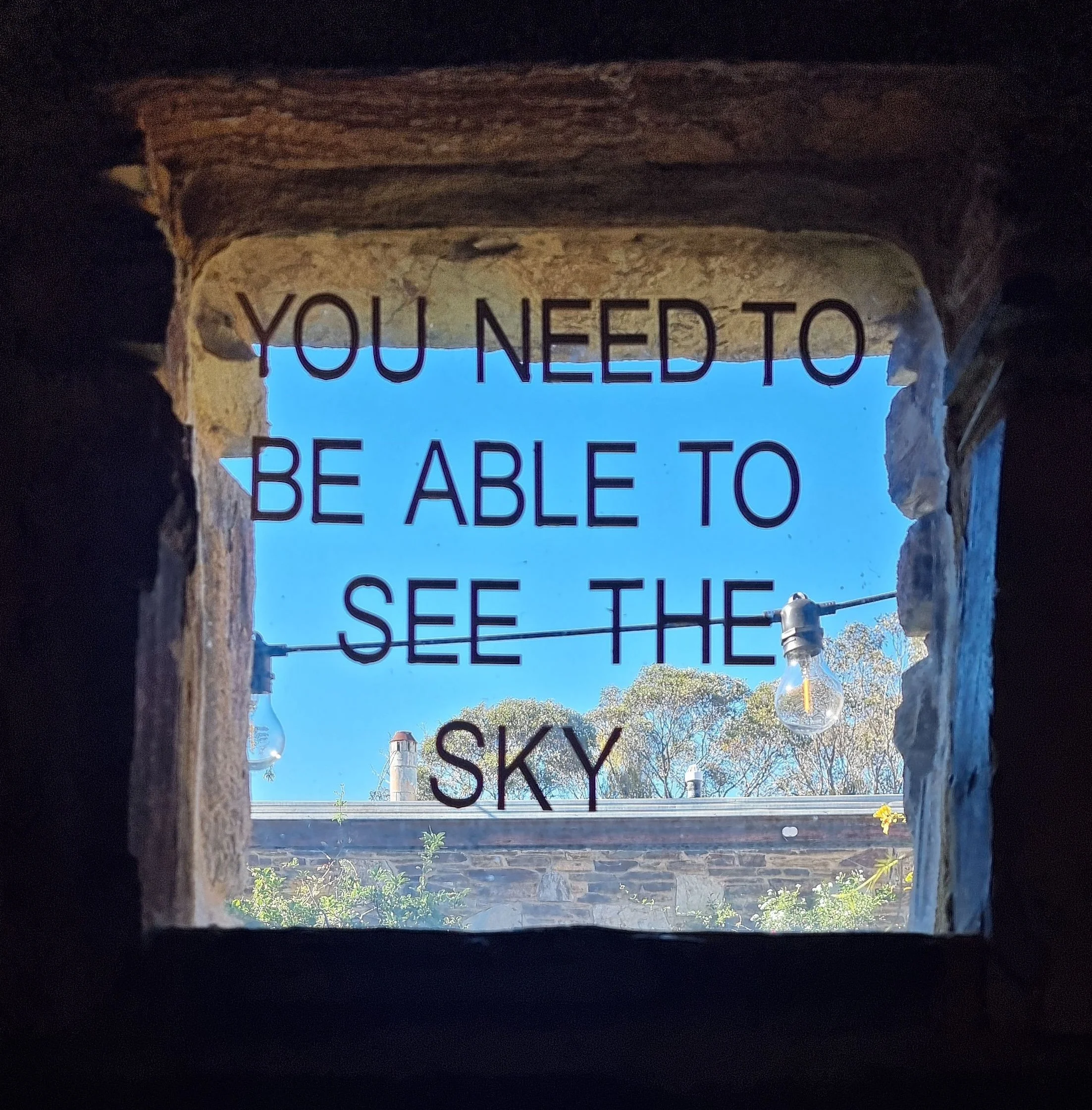 Window lettering reading "You need to be able to see the sky" across a small stone window, with blue sky and gum trees beyond