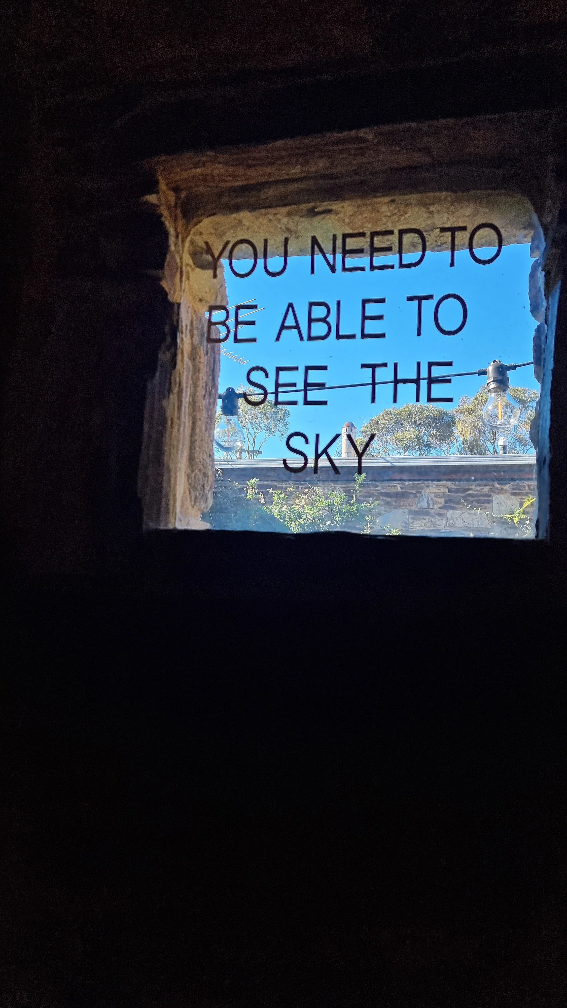 Window lettering reading "You need to be able to see the sky" across a small stone window, with blue sky and gum trees beyond