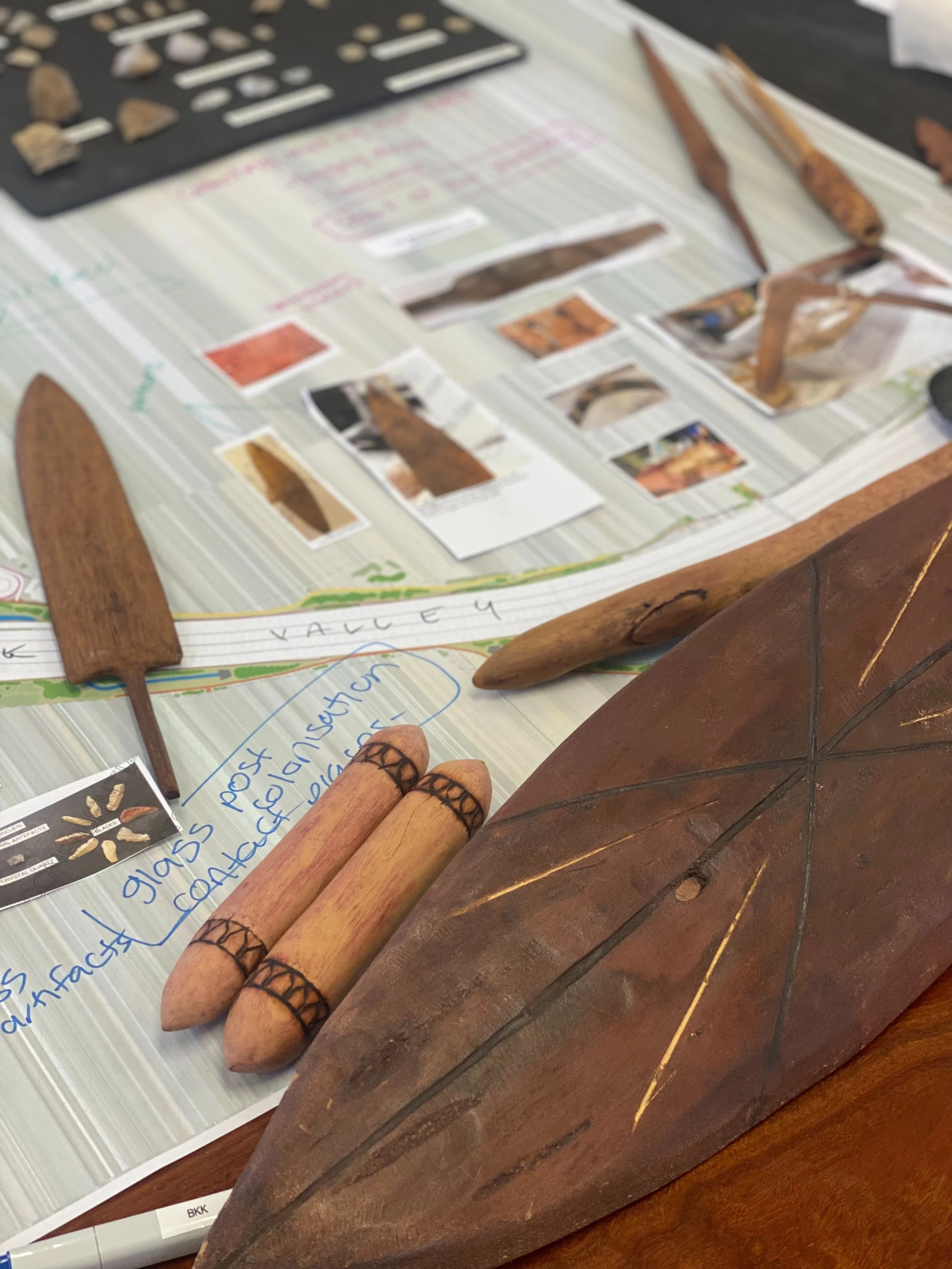 Carved timber cultural objects laid on a table with printed reference images and handwritten notes, part of the design process