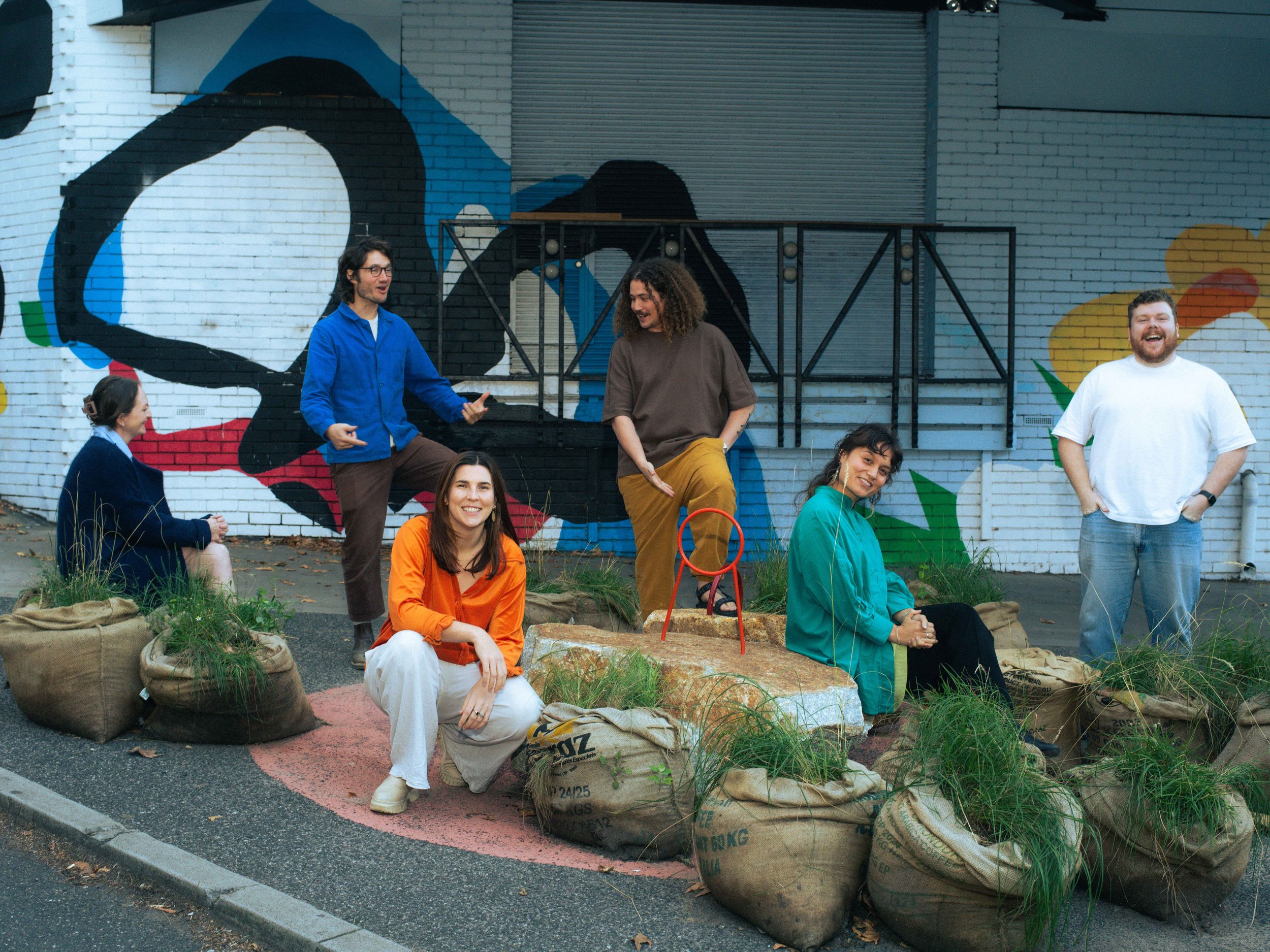 The Winsor Kerr team laughing and chatting among the hessian sacks and grasses of PARKing, in front of a painted mural in Collingwood