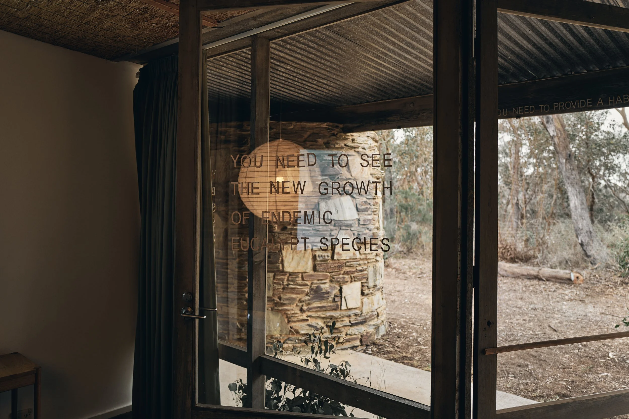 View from inside through glass lettered "You need to see the new growth of endemic eucalypt species", looking out to a stone chimney and bushland