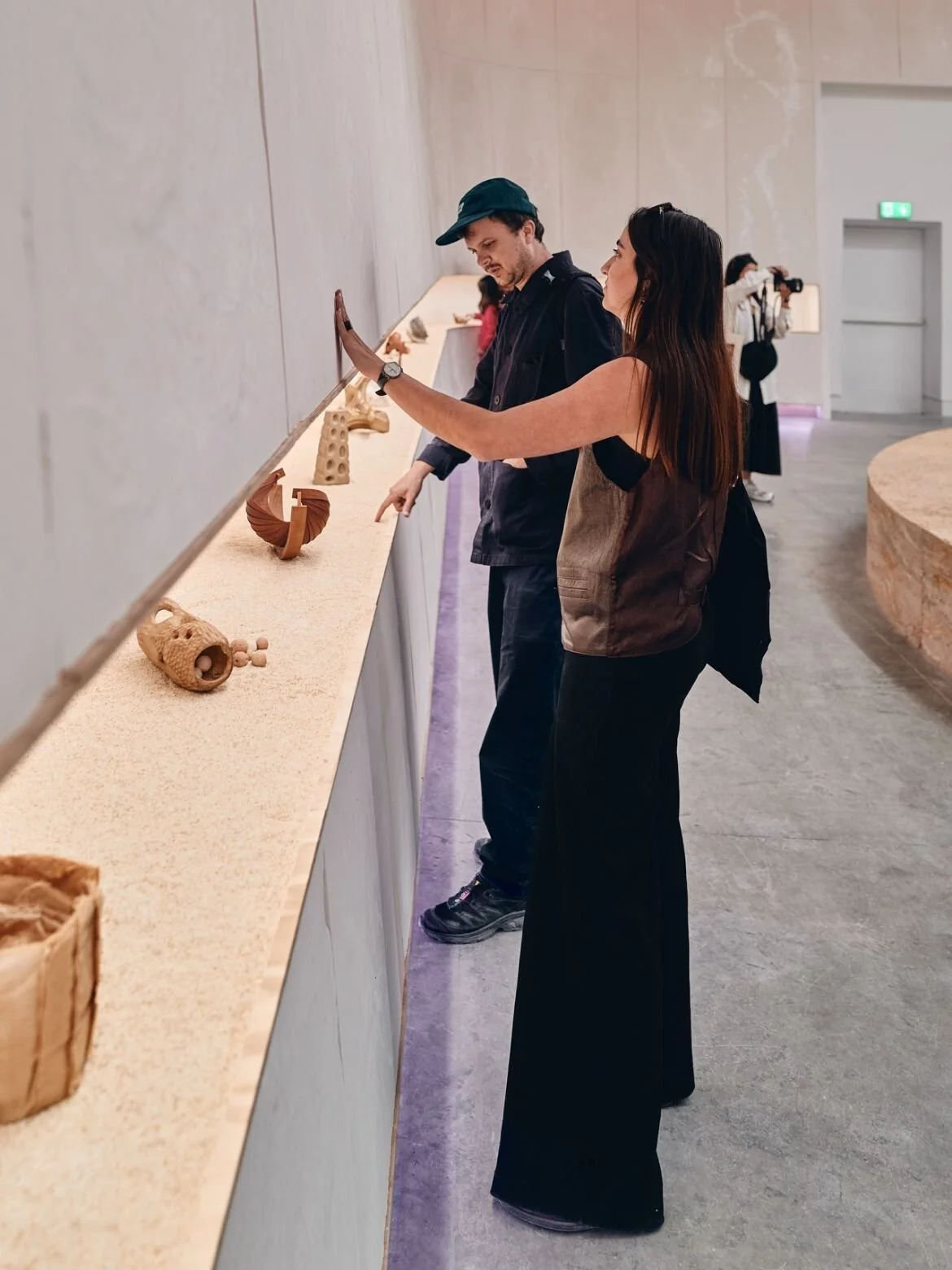 Two visitors looking at small objects of home made by Australian architecture students, displayed along a long shelf