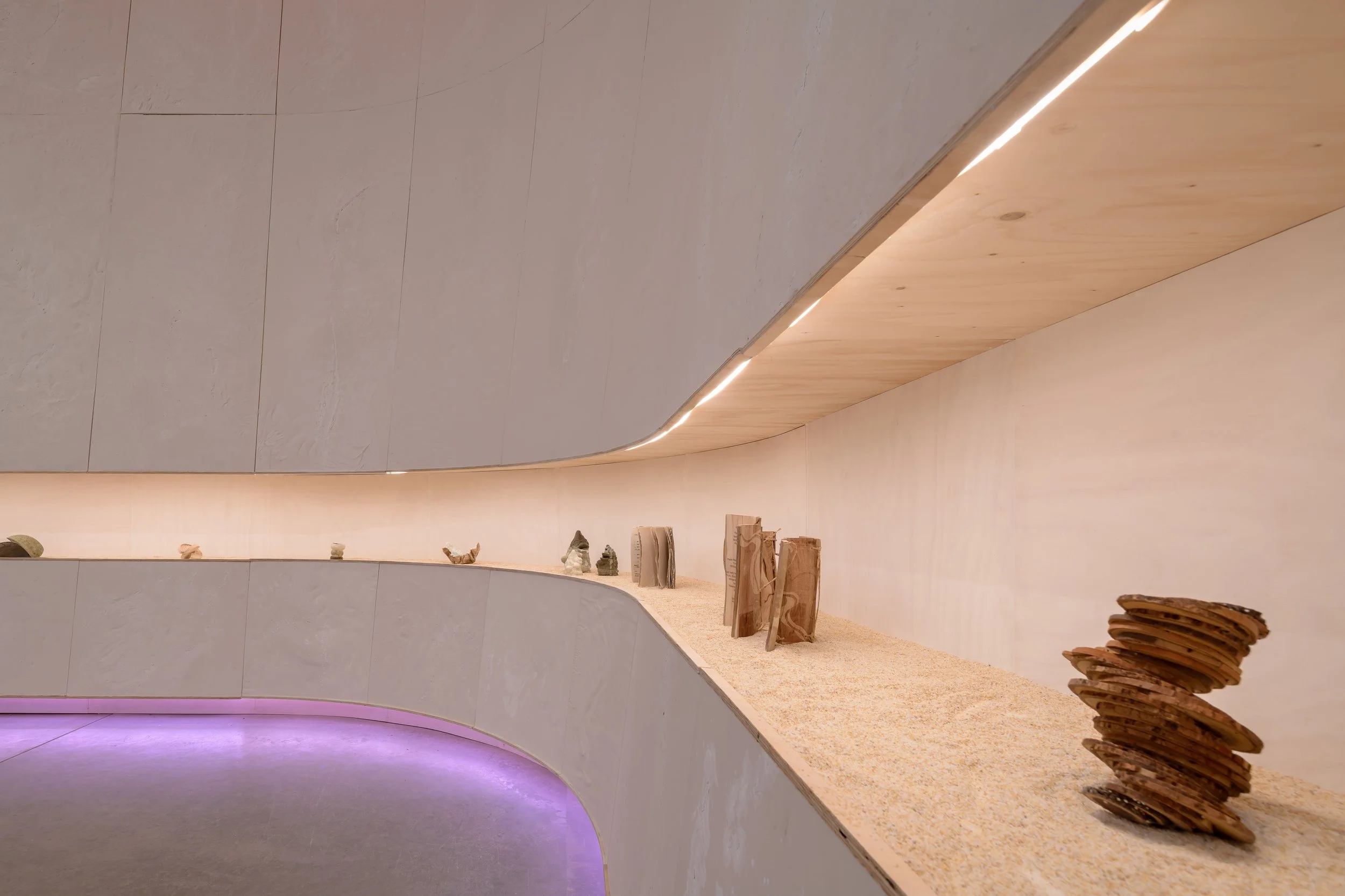 Long curved shelf of objects wrapping around the pavilion above the sand floor, lit with soft purple light