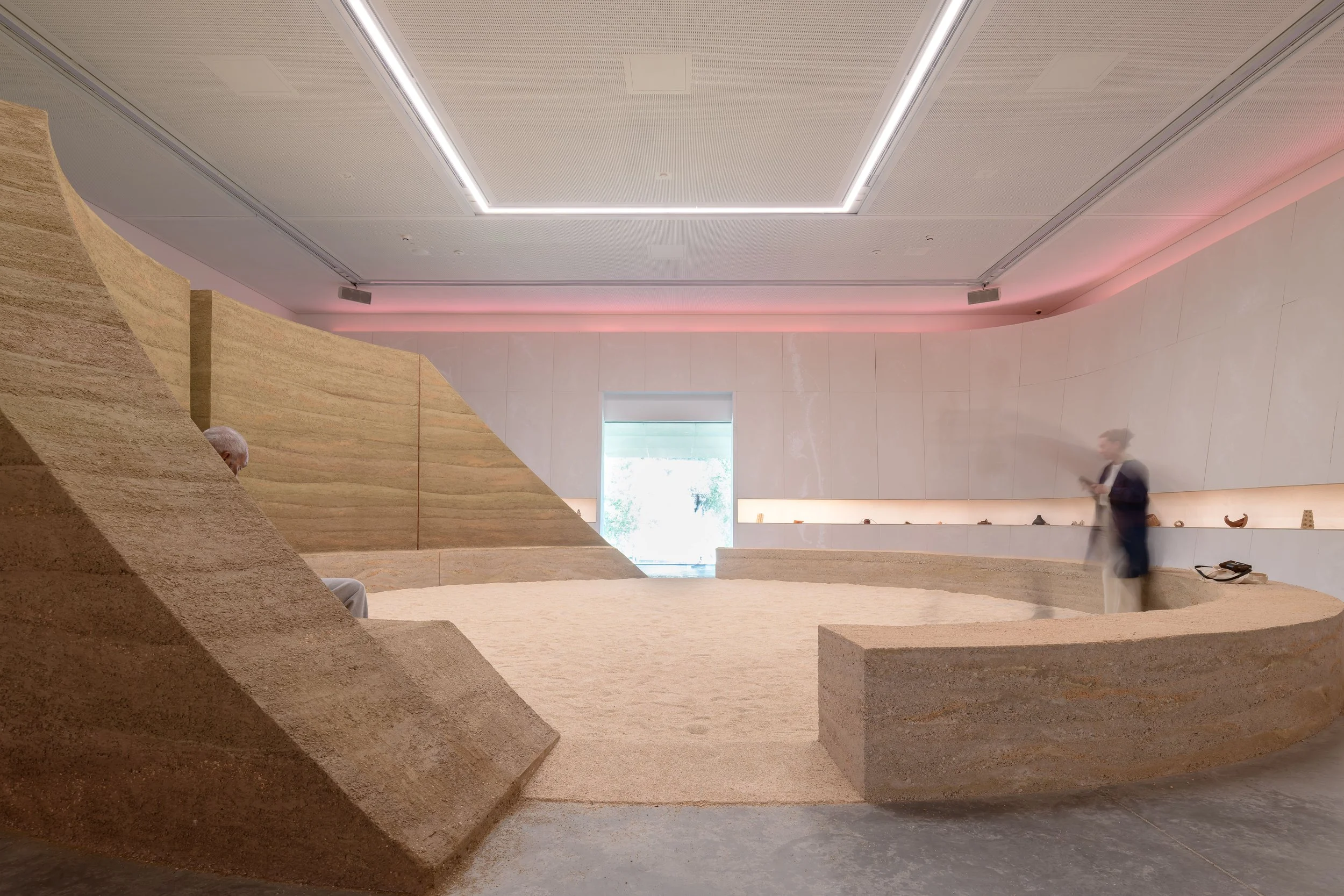 Pavilion interior with rammed earth walls, a sand floor and scattered earth blocks beneath a patterned ceiling