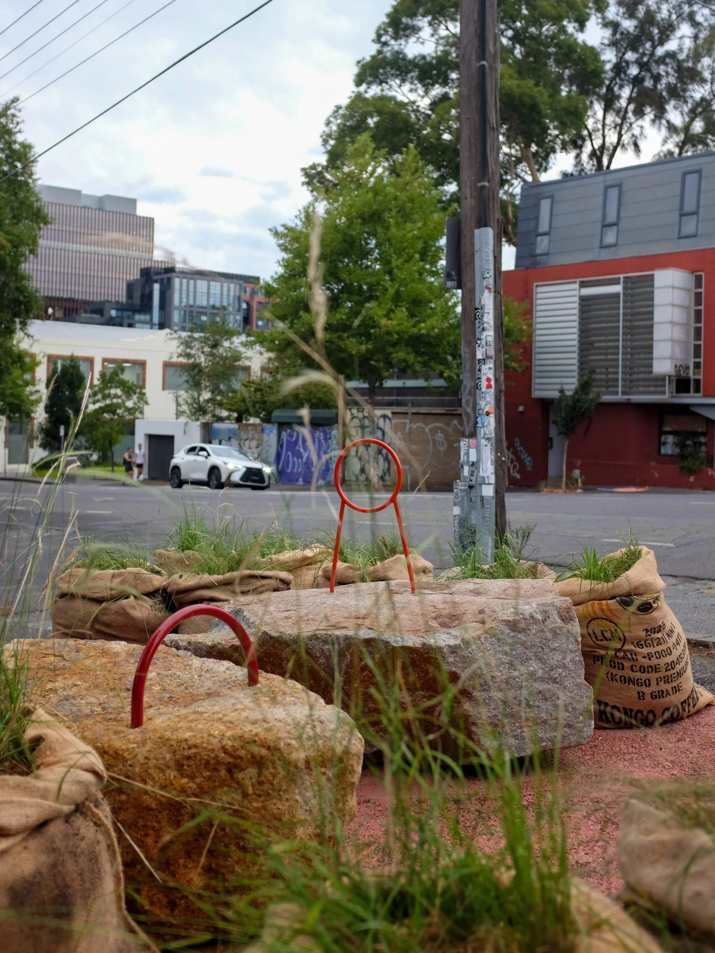 Hessian sacks, granite blocks and red steel loops set into the parking space, with the street's buildings and trees behind