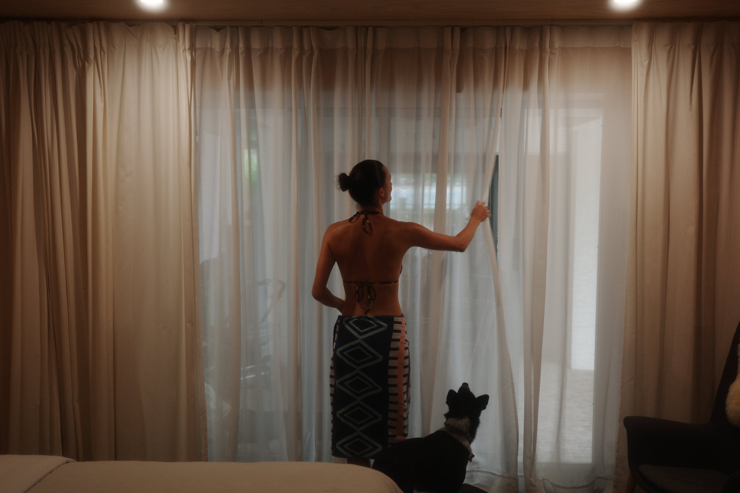 A person drawing back sheer curtains in the bedroom, with a dog sitting beside them