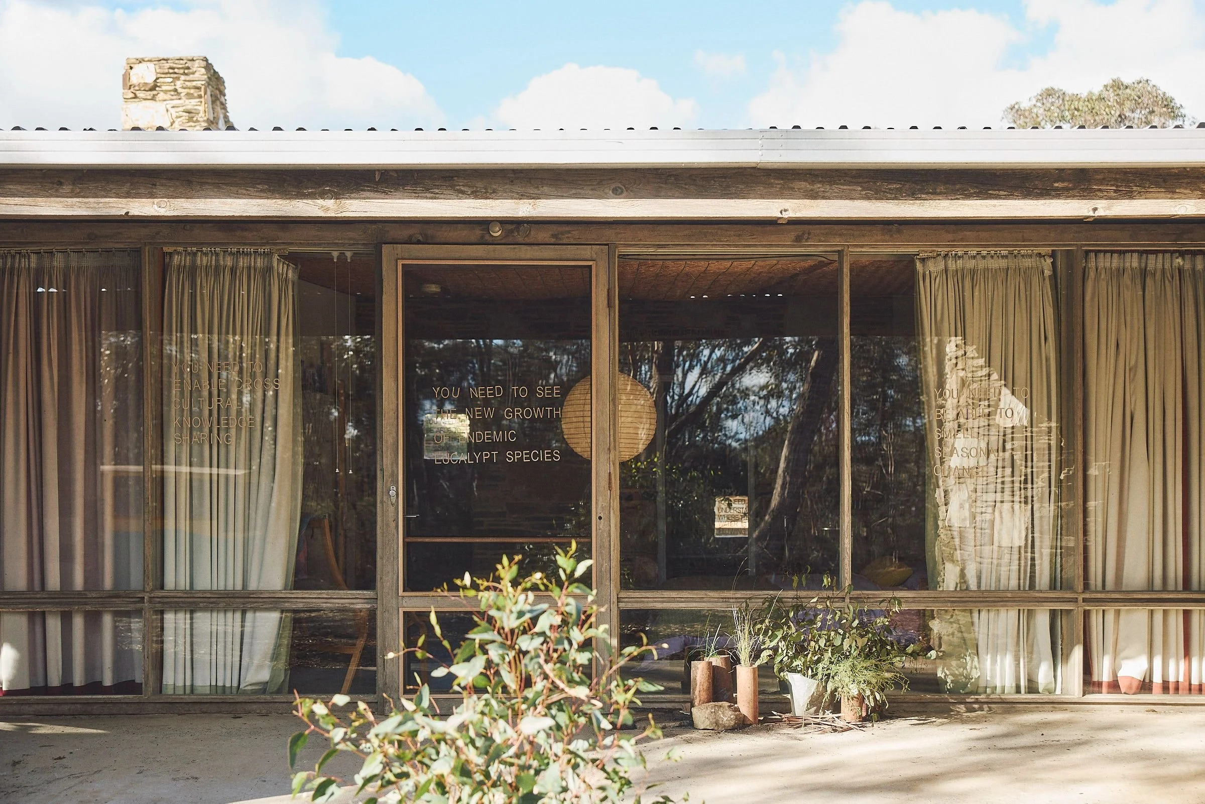 Timber and glass front of Boyd Baker House, with door lettering reading "You need to see the new growth of endemic eucalypt species" and potted plants outside