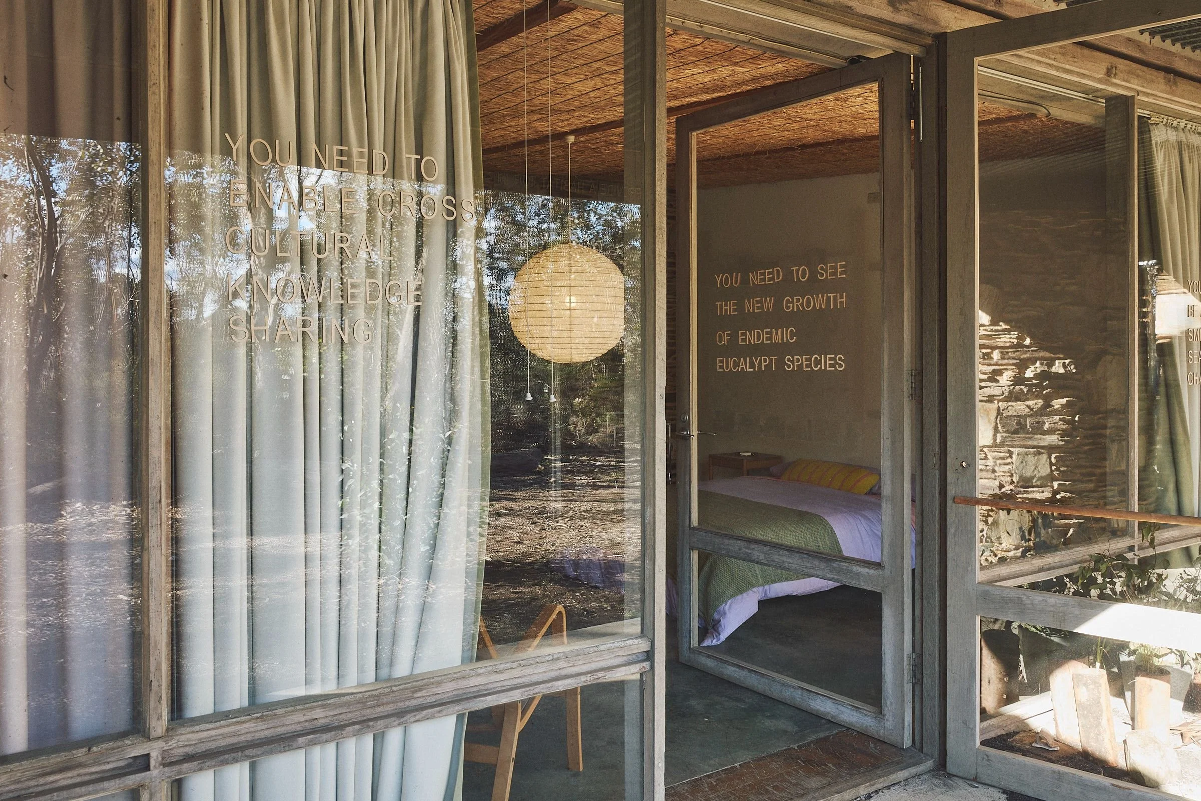 View through a lettered glass door into a bedroom, with linen curtains and a paper lantern