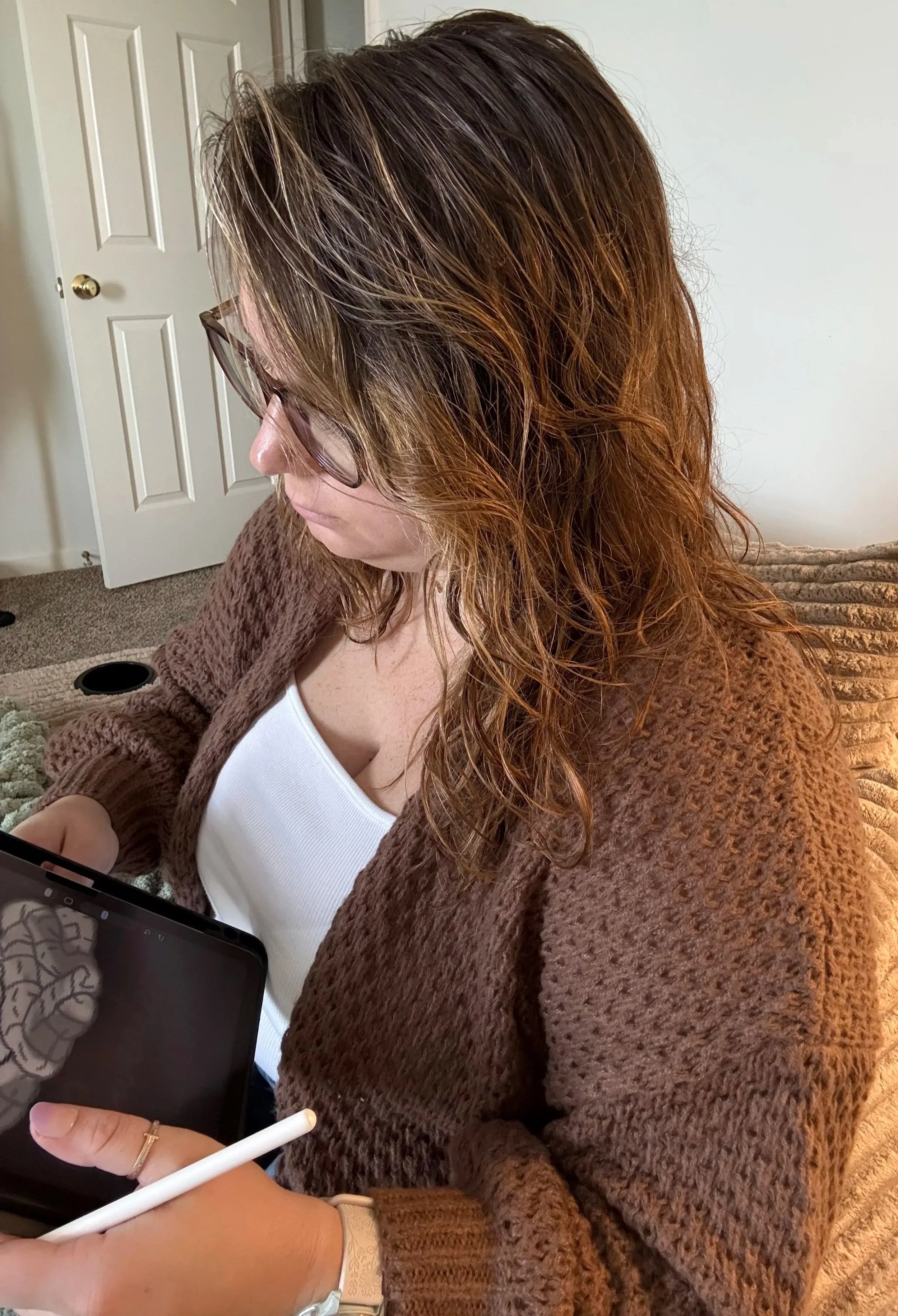 A woman with long, wavy, brown hair with blonde highlights, wearing glasses and a brown knitted cardigan, sitting on a beige couch, holding a tablet, and using a white stylus.
