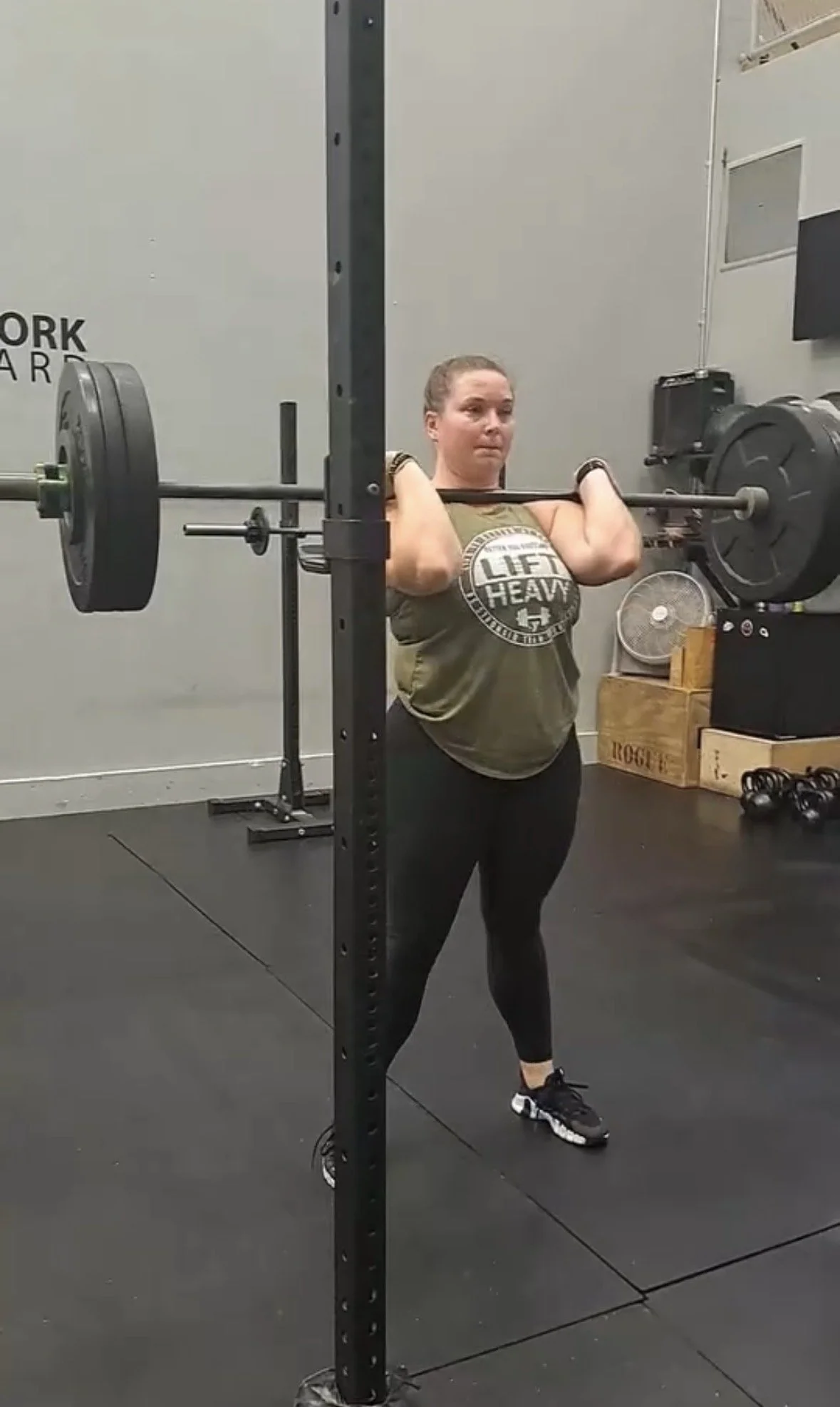 A woman lifting a barbell during a workout at a gym, standing inside a squat rack with weights.