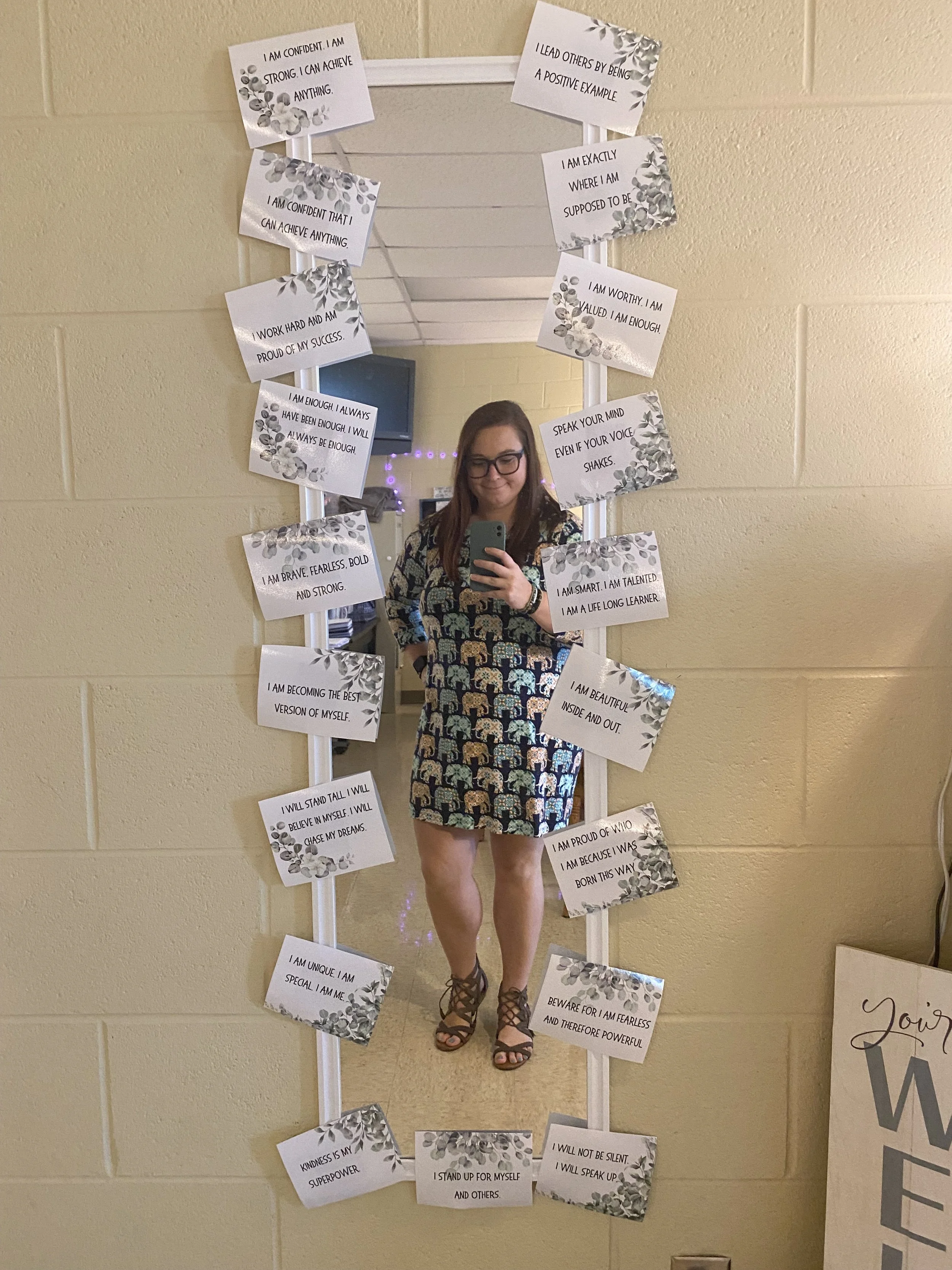 A woman taking a selfie in front of a large mirror decorated with inspirational messages on paper. The woman is wearing a dress with an elephant pattern and sandals. The motivational notes are surrounded by leaf illustrations and include affirmations such as, "I am confident I can achieve anything," "Speak your mind even if your voice shakes," and "Kindness is my superpower." The background includes a yellow wall, a small blue monitor, and some purple lights.
