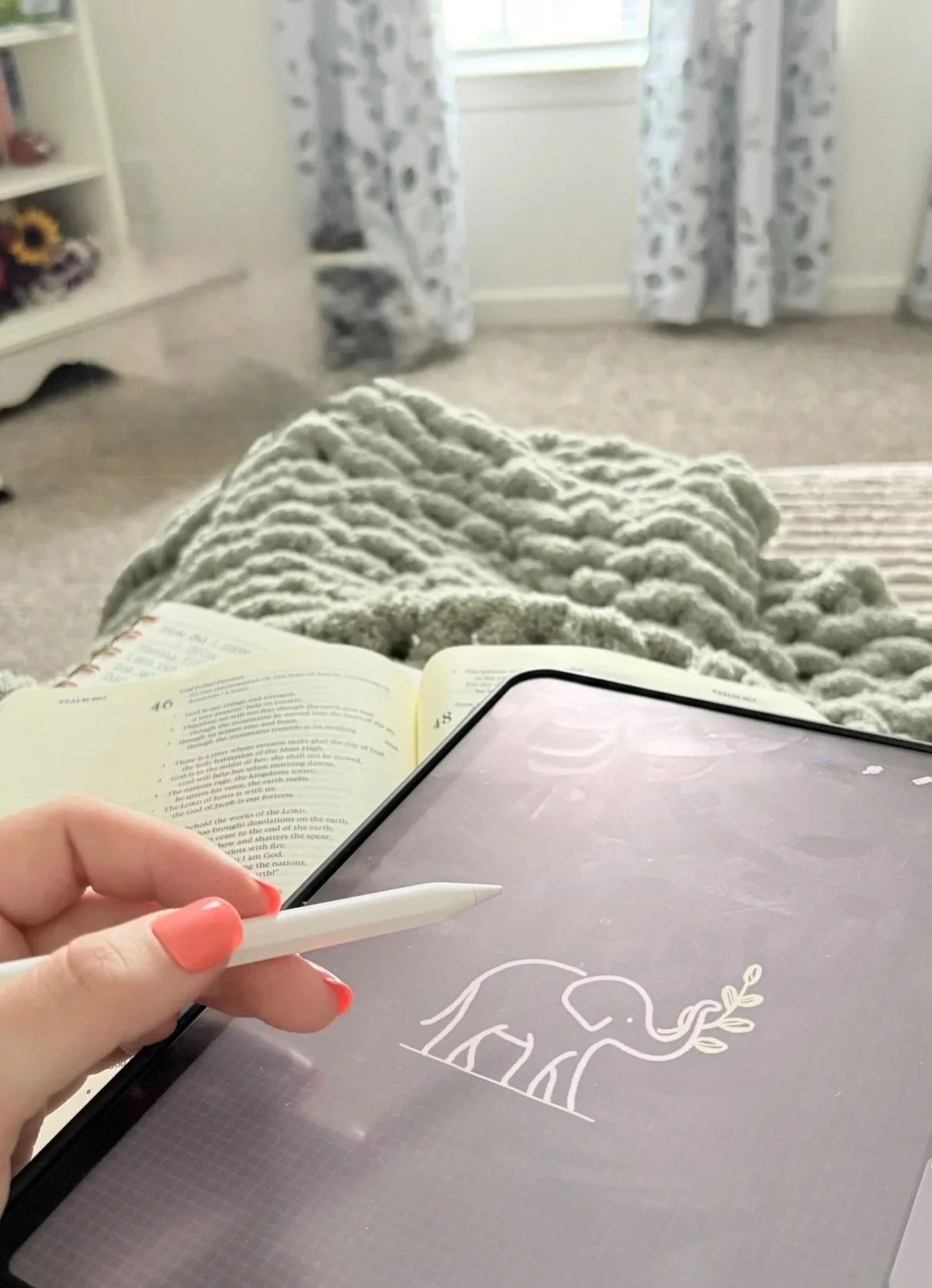 A person holding a stylus over a tablet displaying a line drawing of an elephant with leaves, while sitting on a bed or couch covered with a plush, textured blanket, with an open book nearby. In the background, there are floral curtains and a window.