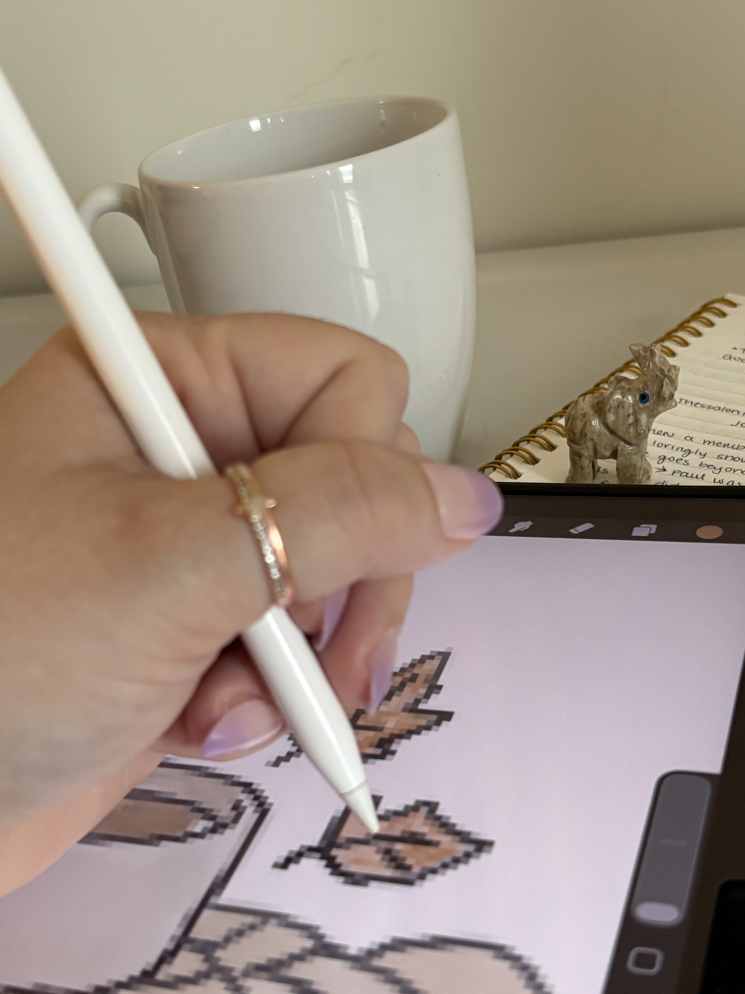 Person holding a white stylus writing on a pixelated digital drawing of a dragon on a phone screen, with white mug and notebook in background.