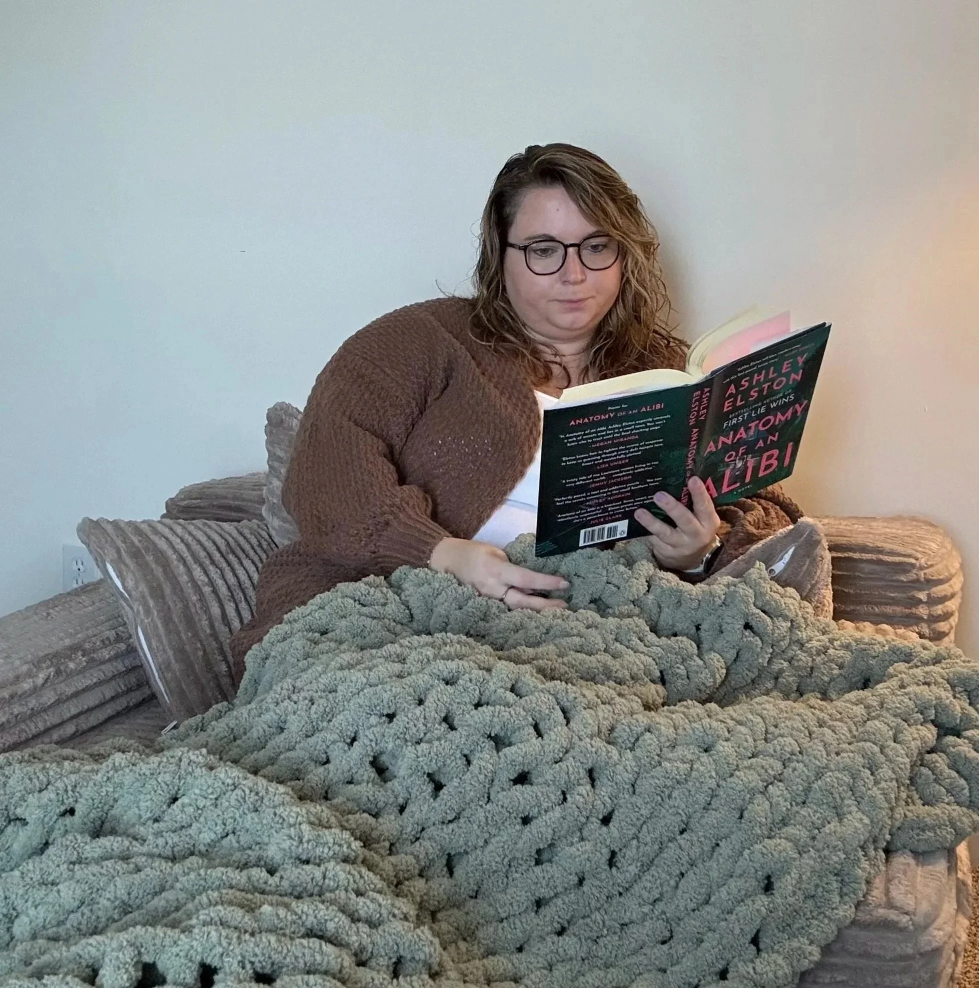 A woman with glasses and wavy hair is sitting on a bed, reading a book titled 'Anatomy of an Alibi' in a room with plain white walls and a beige blanket.
