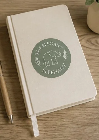 A white hardcover notebook with a green circular logo featuring an elephant and the words 'The Elegant Elephant' on a wooden surface, with a pen and a potted plant nearby.