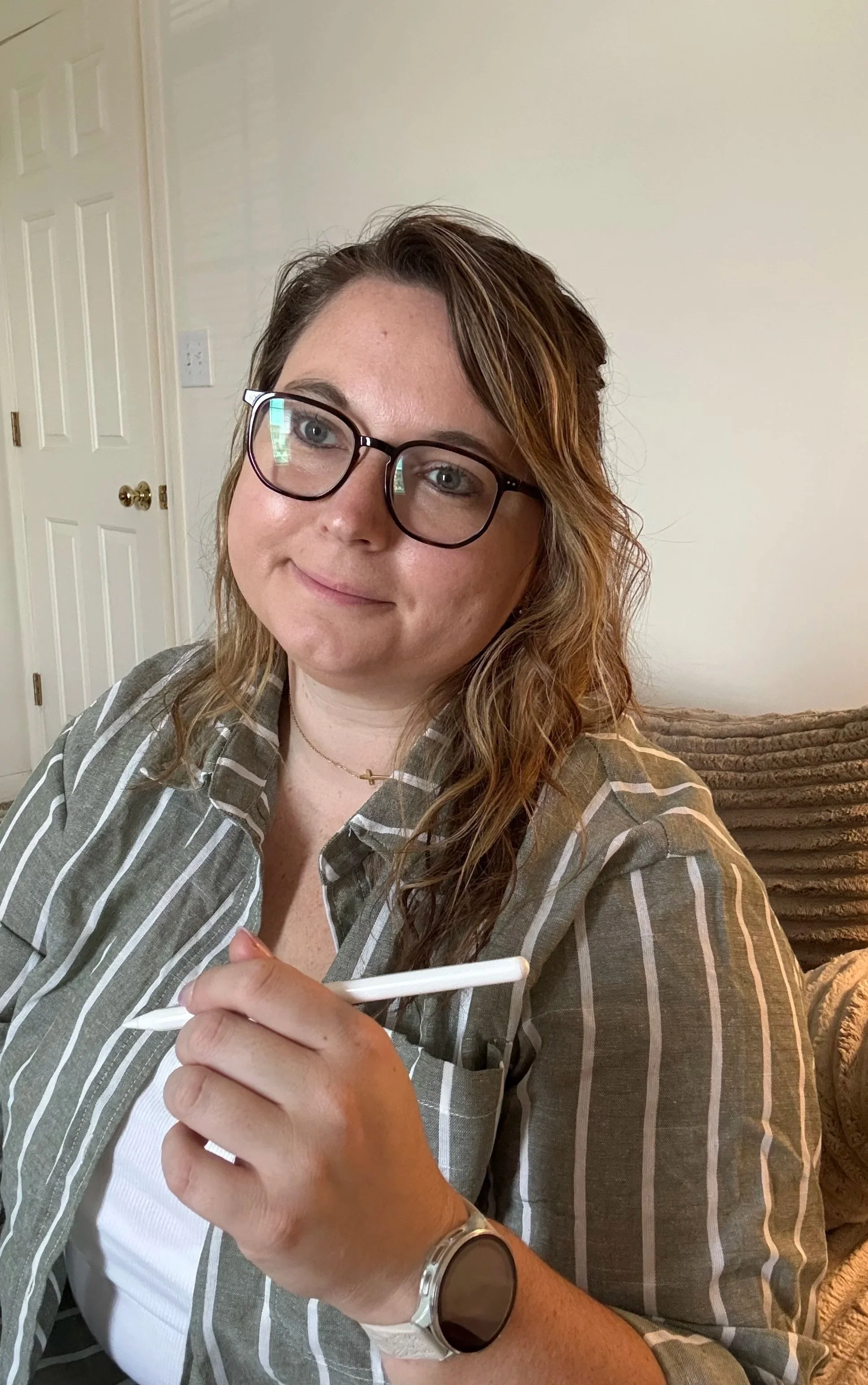 A woman with glasses and wavy brown hair sitting on a couch, holding a white stylus in her right hand, wearing a striped shirt and a watch.