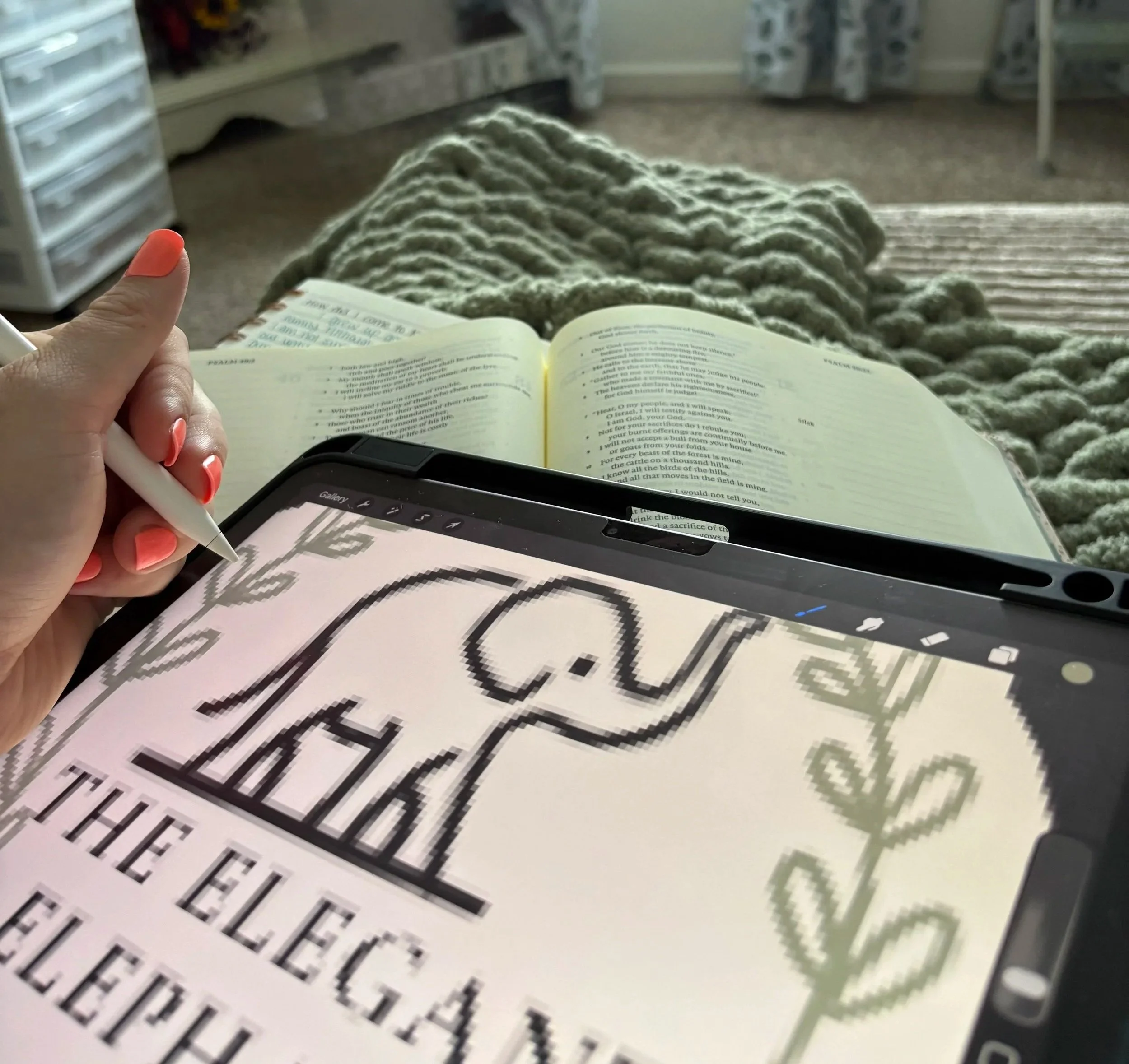 A person holding a white stylus in front of an open Bible with text and highlights, sitting on a bed with a chunky knit blanket, a tablet displaying a black and white image, and a large printed text on paper nearby in a cozy room.