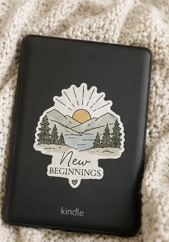 Kindle e-reader with a sticker showing mountains, trees, a river, and a sunrise, with the words 'New Beginnings'.