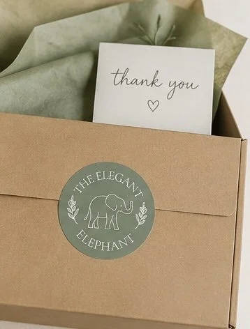 A thank you card with a heart symbol, placed inside a box with green tissue paper and a sticker labeled 'The Elegant Elephant' featuring an elephant illustration.