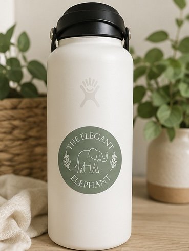 White water bottle with a black lid, featuring a circular logo with an elephant and the words 'The Elegant Elephant' on a wooden table with potted plants in the background.