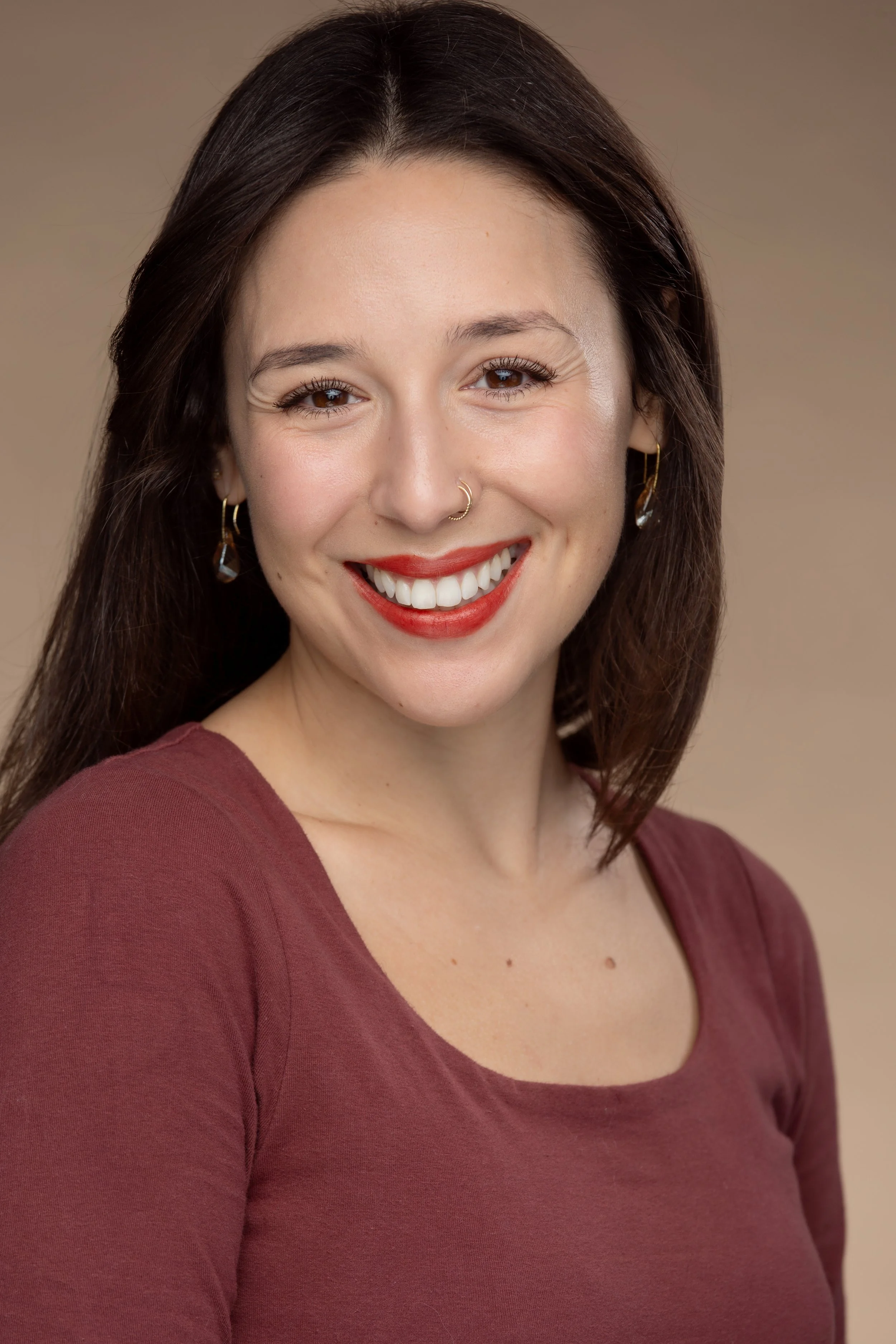 Close-up portrait of a smiling woman with brown hair, wearing a maroon top and gold earrings, against a beige background.