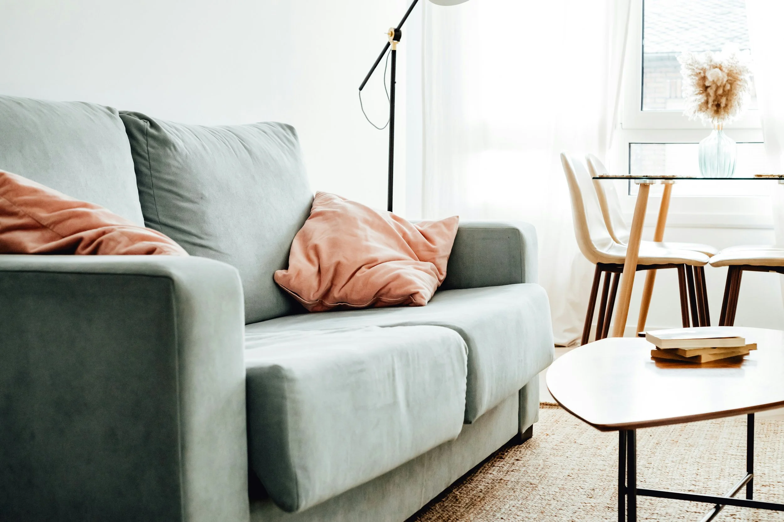 Living room with a light green sofa, pink blankets, a coffee table with books, a dining table with wooden chairs, a tall vase with fluffy white and brown flowers, a large window with curtains, and a floor lamp.