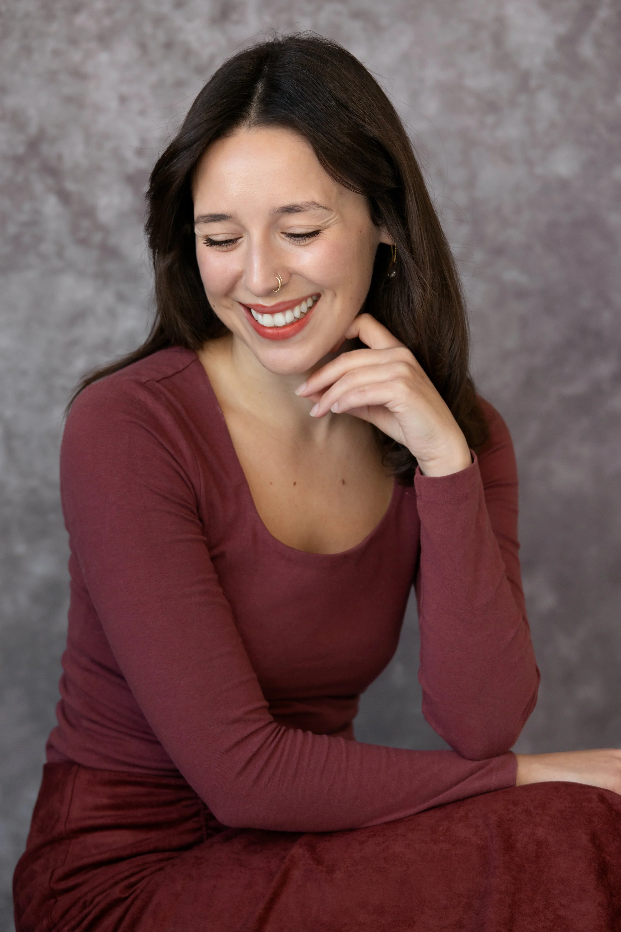 A young woman with long dark hair, wearing a maroon long-sleeve shirt, is smiling and looking downward with her eyes closed. She has a nose ring and an earring, and is posing with her hand gently touching her chin, against a textured gray background.