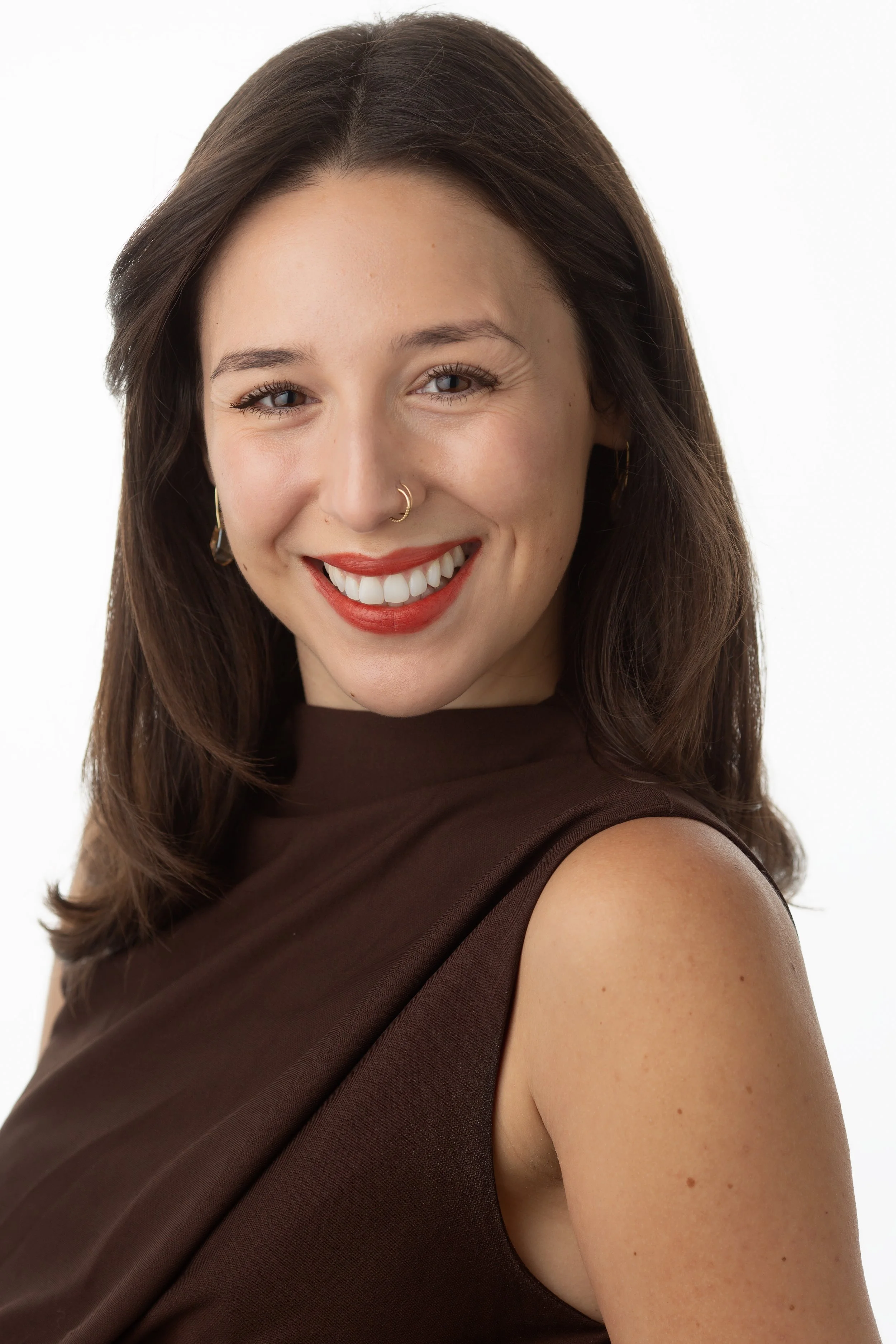 Close-up portrait of a smiling woman with shoulder-length brown hair, wearing a sleeveless dark brown dress, gold hoop earrings, and a gold nose ring, against a plain white background.