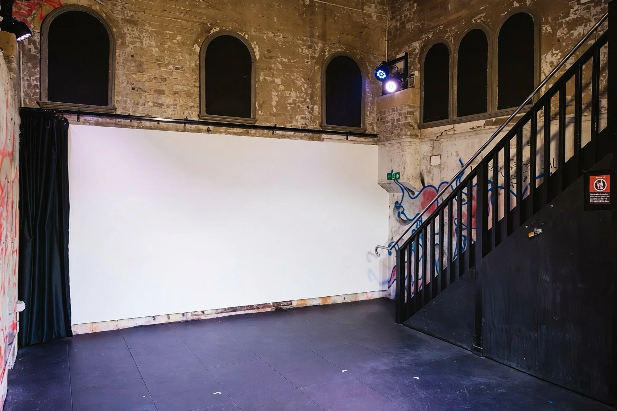An indoor space with a brick wall and arched windows. There is a white projection screen, black curtain, colorful graffiti on the wall, and a staircase with a black railing. Two stage lights are mounted on the wall, casting blue and purple light.