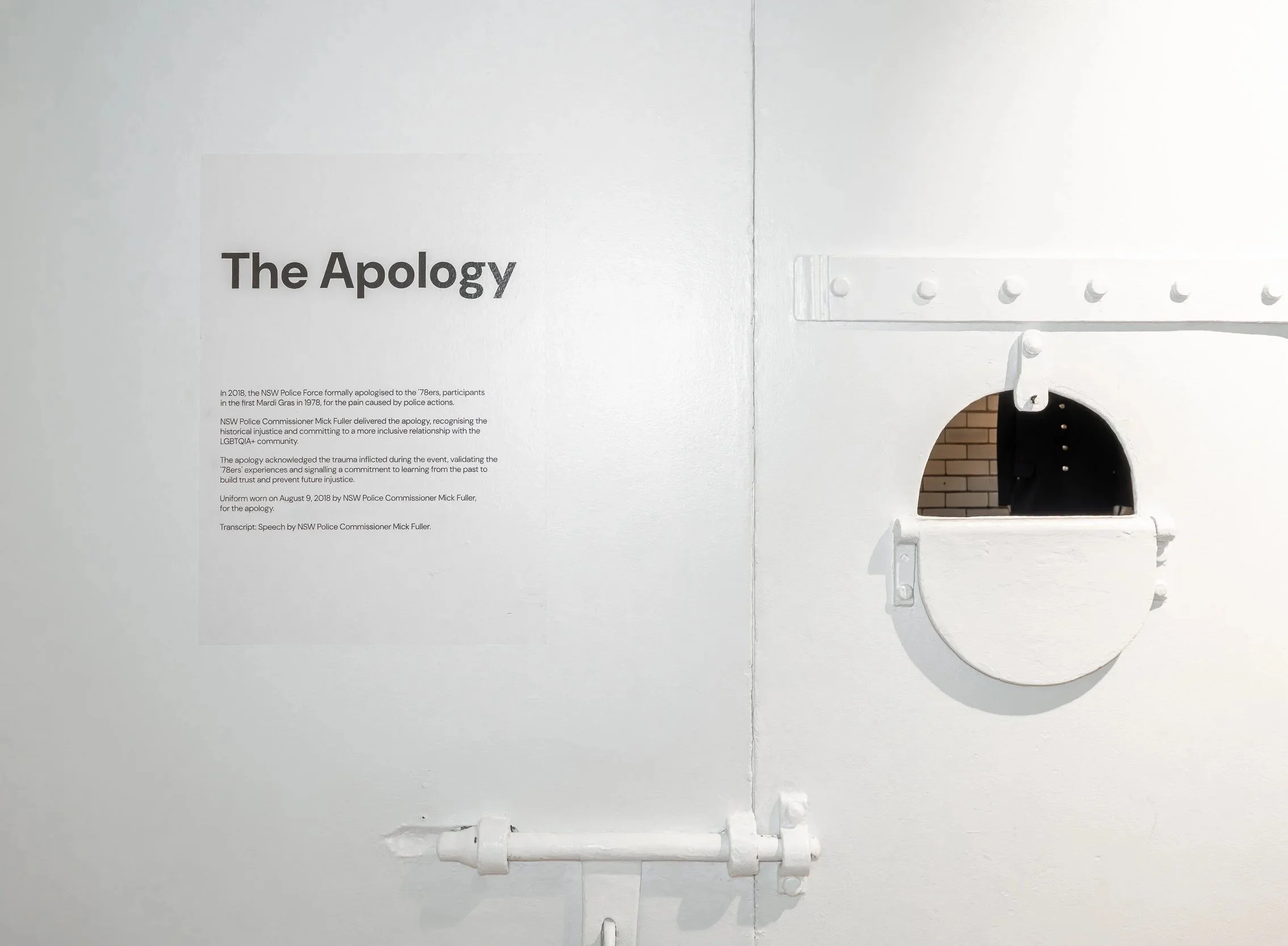 A white wall with black text that reads 'The Apology' and a paragraph of smaller text below. To the right, a white metal structure with bolts and pipes, with part of a brick wall and a dark opening visible.