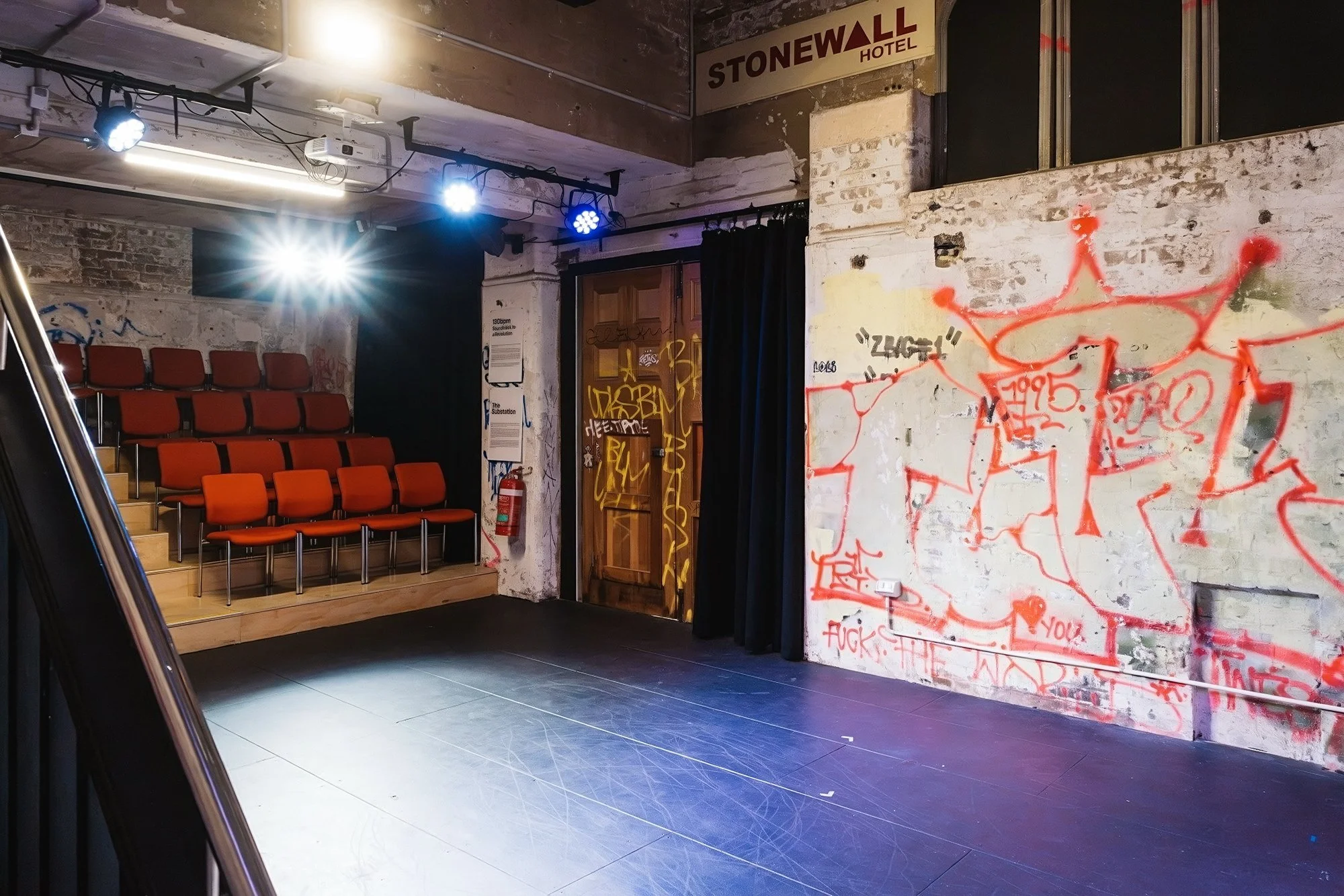 An indoor space with red upholstered chairs arranged in rows, a black curtain, and a white brick wall with red graffiti. There is a sign that says 'STONEWALL HOTEL' at the top right. The ceiling has some exposed pipes and lighting fixtures, including blue and white stage lights. A wooden door is visible in the back with graffiti, and a fire extinguisher is mounted beside it.