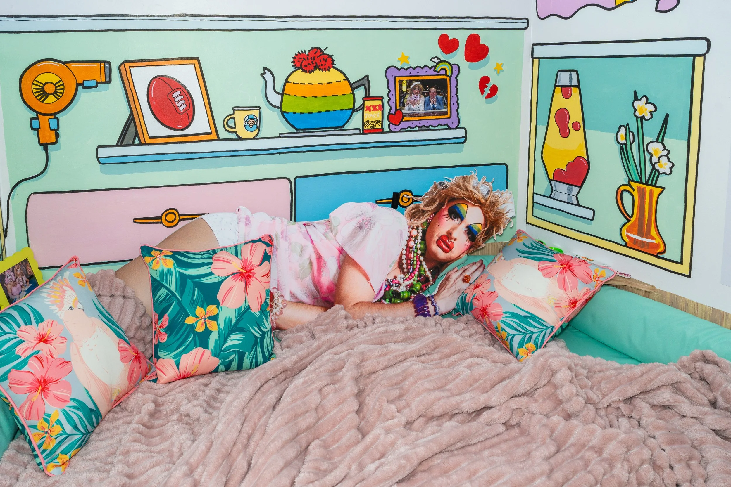 Colorful bedroom with wall art displaying a rainbow teapot, framed photo, lamp, and hearts, featuring a person with vibrant makeup and jewelry lying on a bed with floral pillows and a plush blanket.