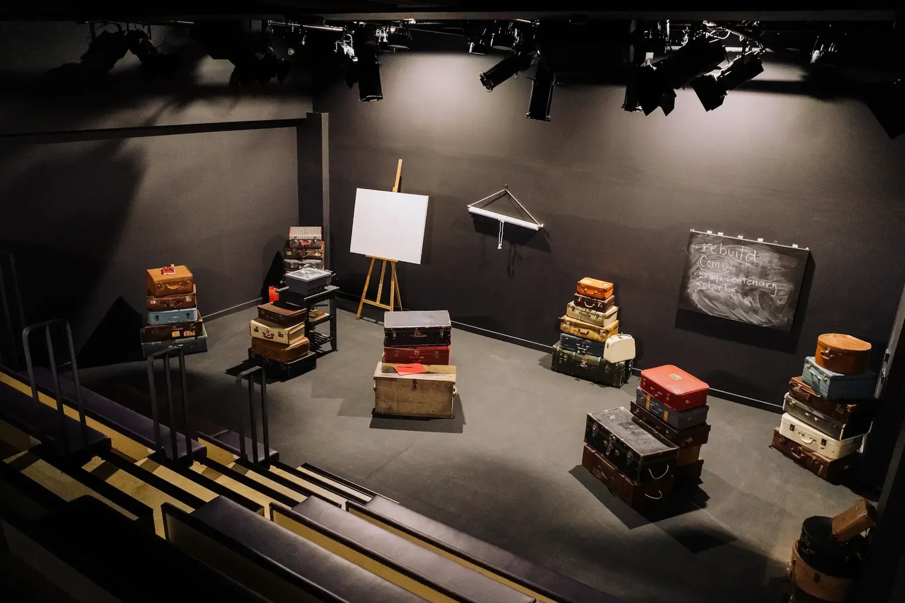 Empty theater stage with stacks of suitcases, a whiteboard on an easel, a chalkboard with writing, and stage lights overhead.