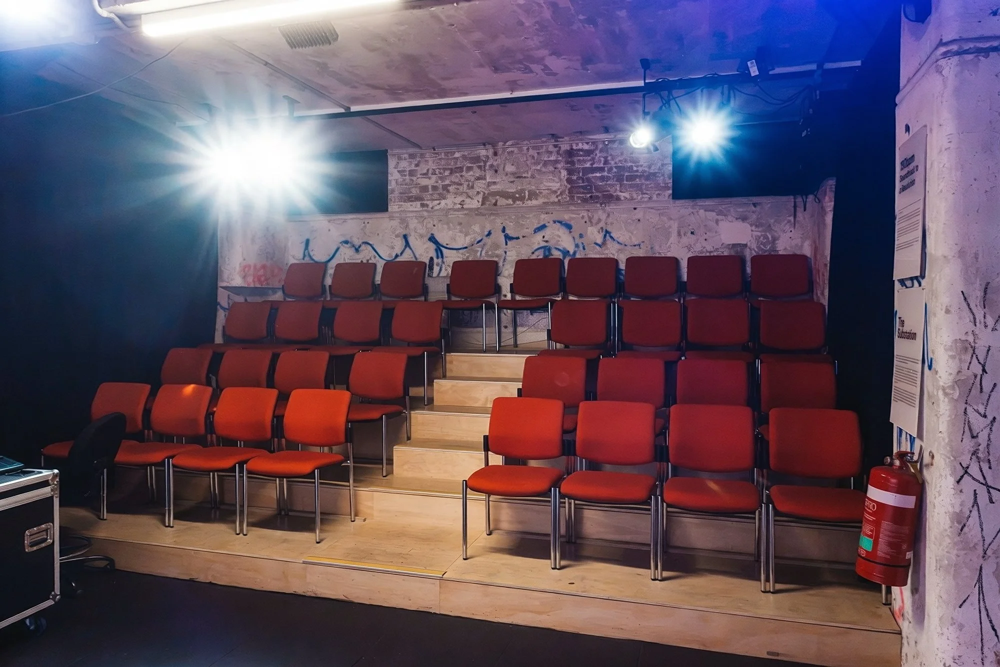 An empty small auditorium or theater with multiple rows of red chairs arranged on a tiered wooden platform. The room has walls with graffiti and a brick section decorated with blue graffiti or designs. Bright lights are mounted on the ceiling, casting light into the room. There is a fire extinguisher at the right corner of the room, and a lectern or equipment case near the bottom left corner.