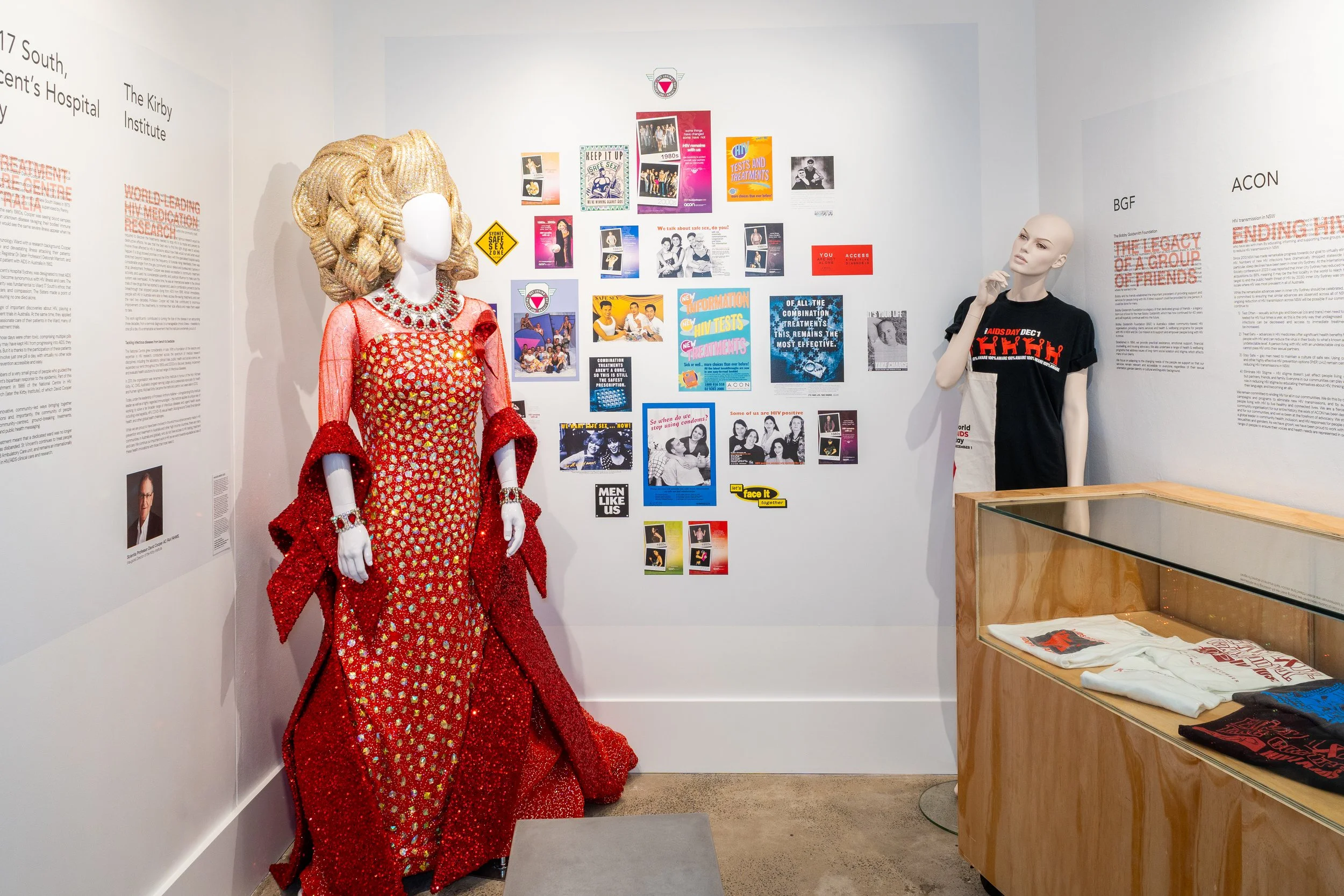 A museum exhibit featuring a mannequin dressed in a red, gold, and pink sequined gown with a large, elaborate wig styled in vintage waves. The background wall is decorated with informational posters and images about HIV/AIDS research and awareness campaigns.