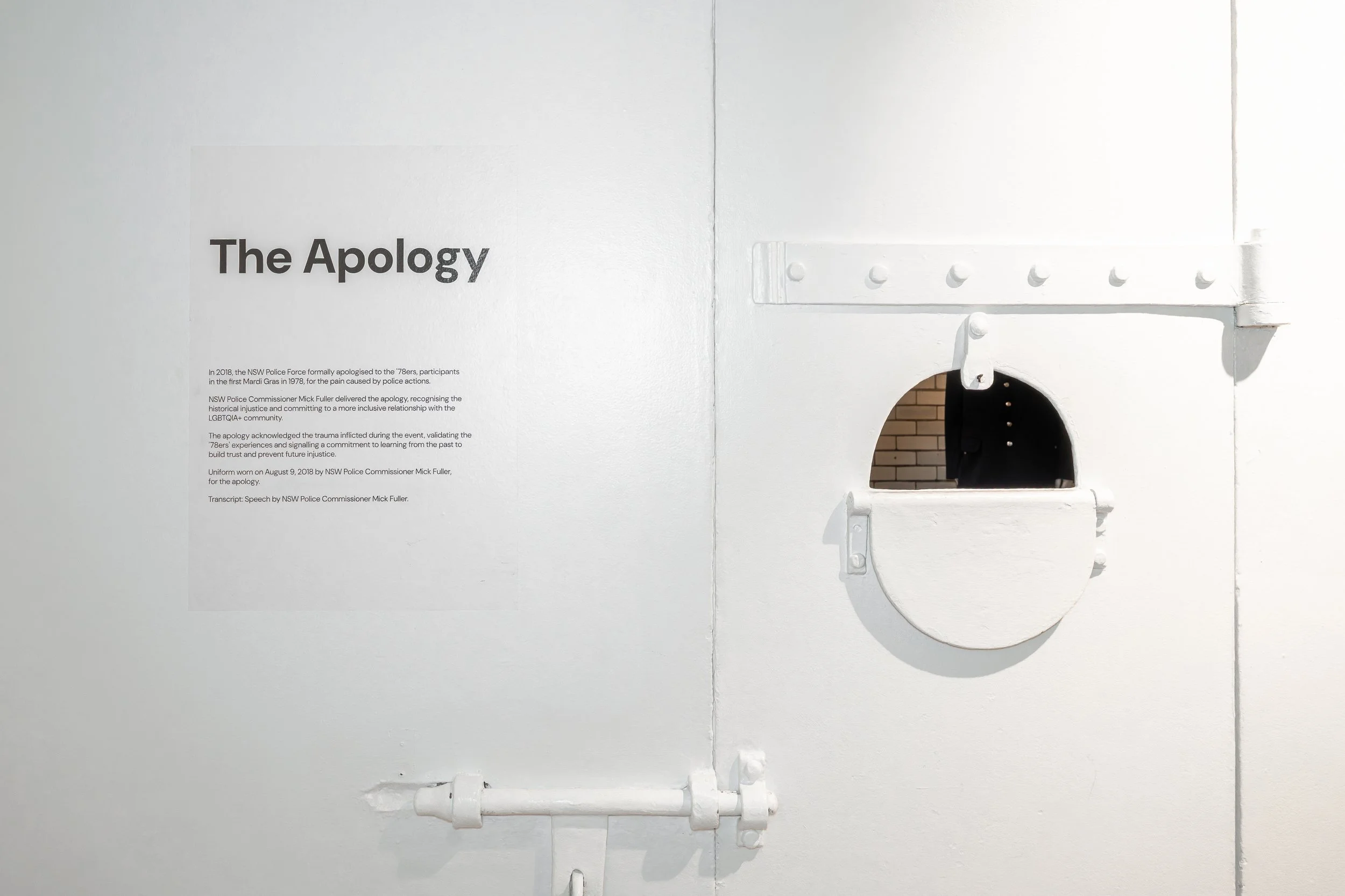 An exhibition display titled 'The Apology' with a white wall in the background. The display includes a paragraph of text discussing an apology by NSW Police Commissioner Mick Fuller in 2018, acknowledging trauma and injustice related to police actions, with a small arched opening below the text showing a person in a dark uniform through the opening.