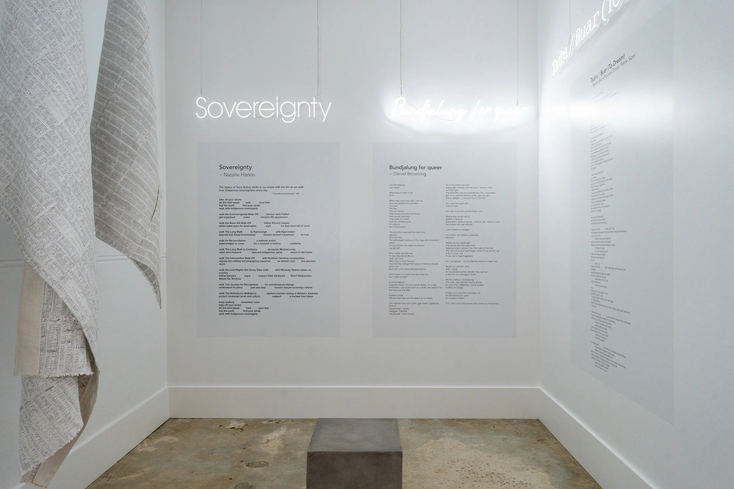 Art gallery exhibit featuring illuminated text on the walls. The words 'Sovereignty' and 'Bundjalung for queer' are visible along with accompanying poetry or descriptions. On the left side, fabric or paper art installations hang, resembling rolled scrolls or scroll-like shapes with written or printed text.