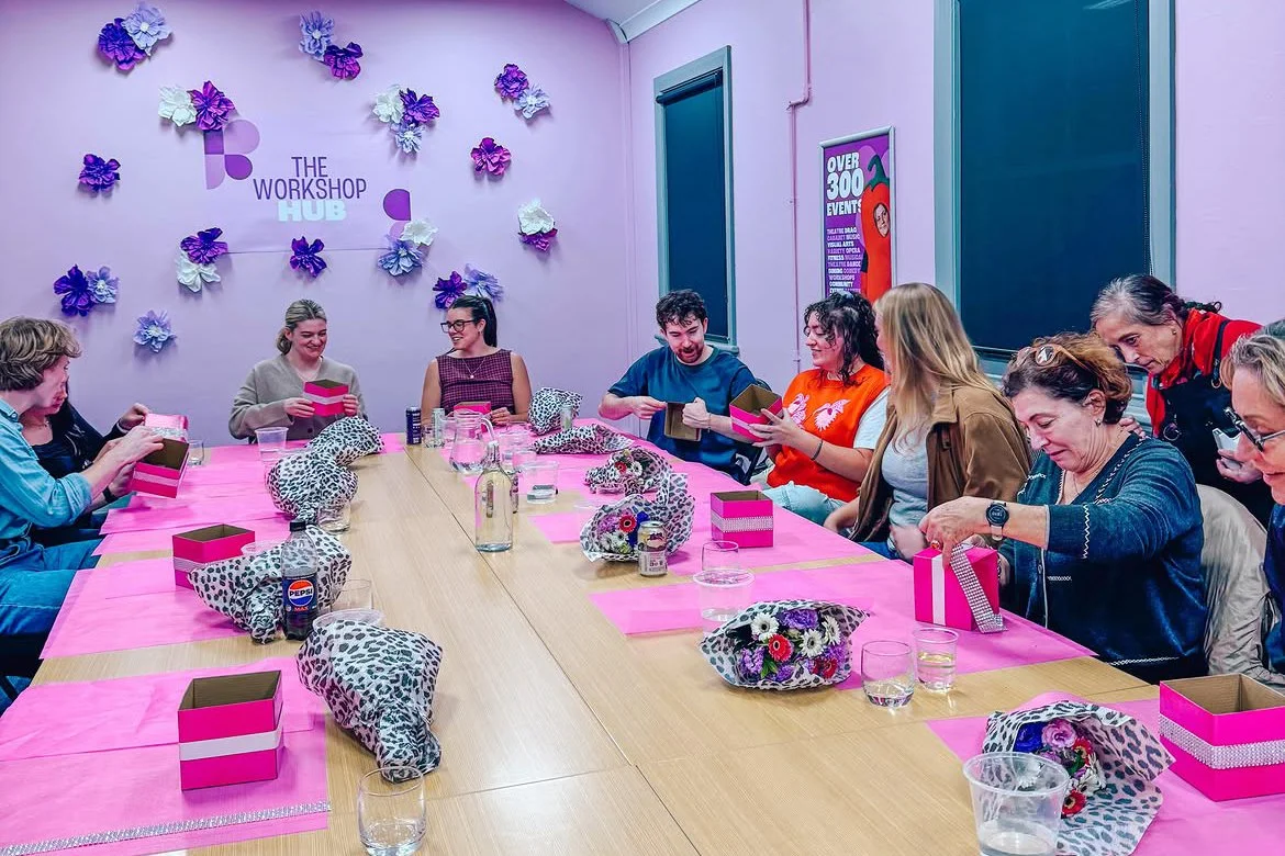 Group of people sitting around a pink table, opening gifts, decorated with leopard print and floral wrapping, in a room with pink walls and floral wall decorations.