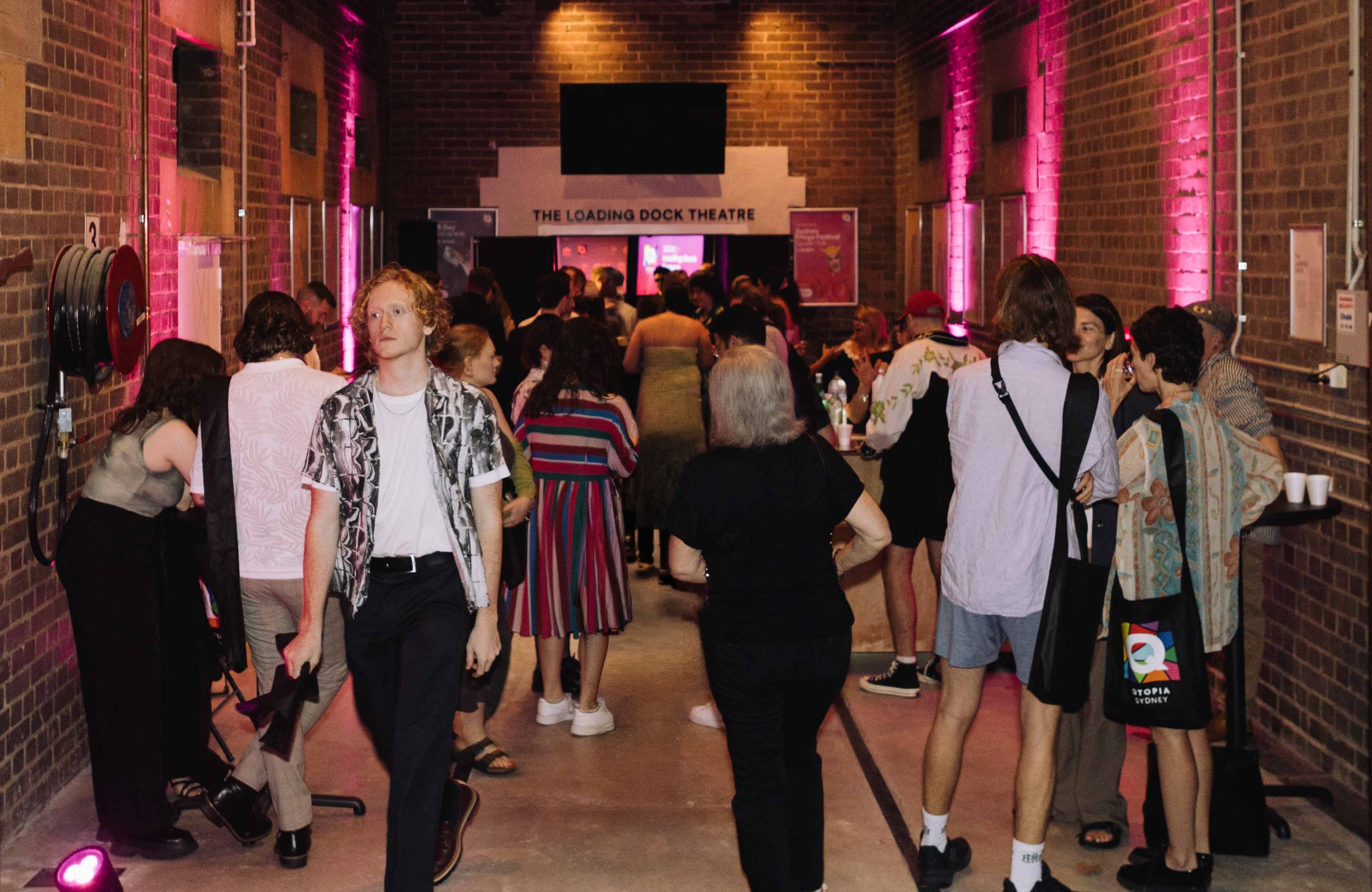 People gathered in a brick-walled indoor venue, with a sign stating 'The Loading Dock Theatre' on the back wall, pink lighting, and large screens.