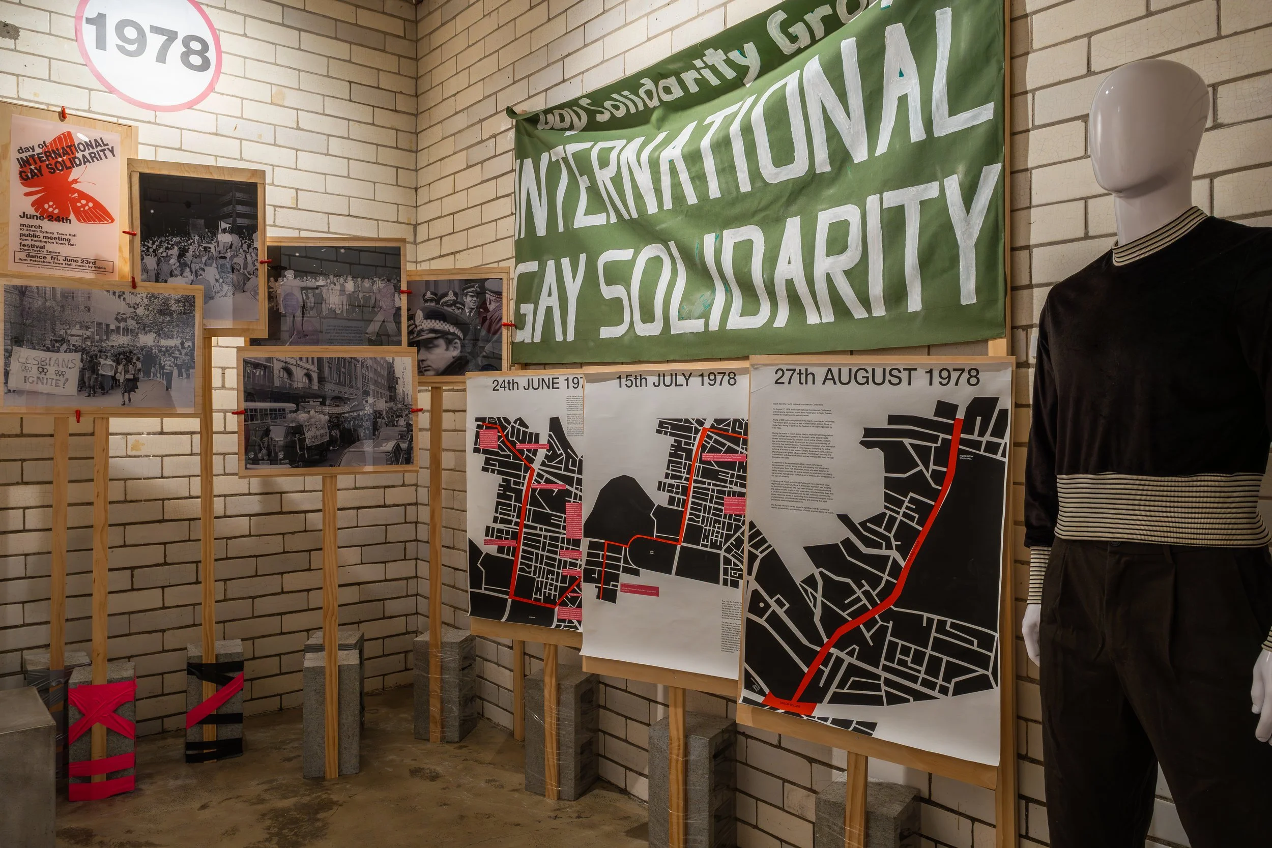 Exhibit with photographs, maps, and banners related to international gay solidarity, including a large green banner with white text, black and white historical photos, and maps of specific dates.