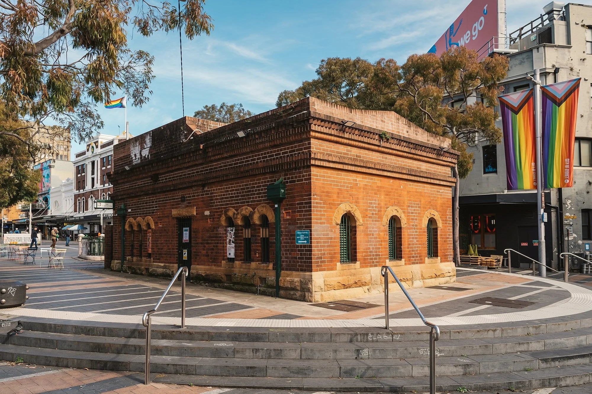 A historic brick building in a city plaza, surrounded by trees and modern buildings, with rainbow flags and a large pink billboard reading 'wego' in the background.